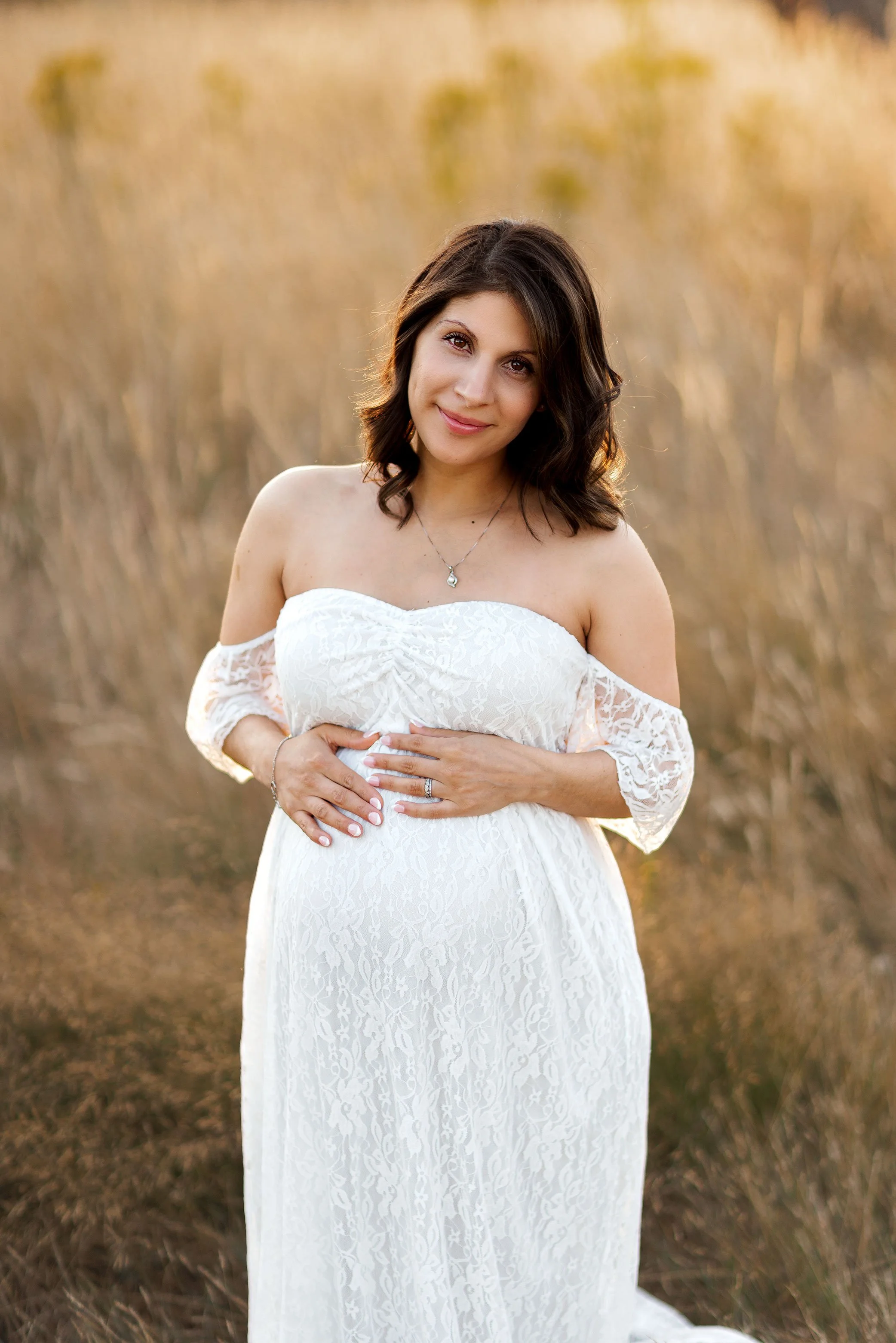 Expecting mother in white lace gown holding baby bump in golden field at sunset.