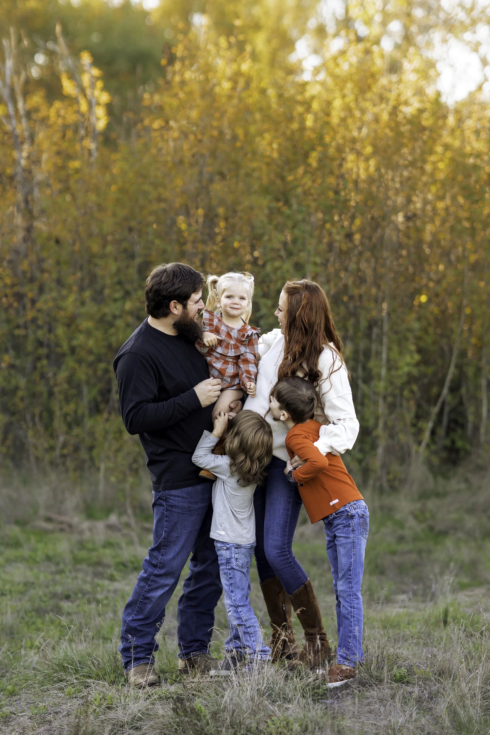 Parents holding toddler while siblings gather close in a grassy field at sunset.