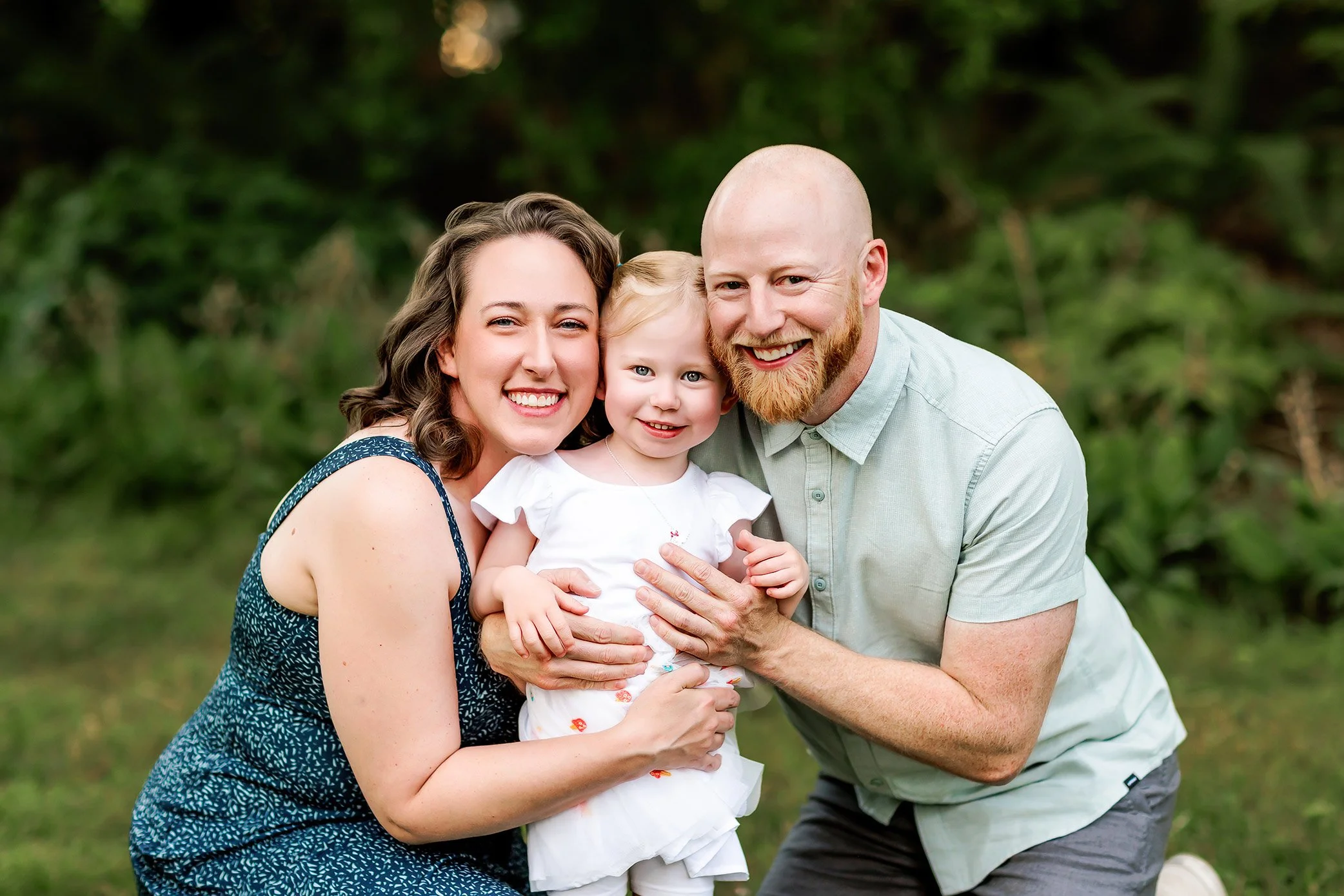 Parents kneeling closely around toddler outdoors, creating connected family portrait at golden hour.