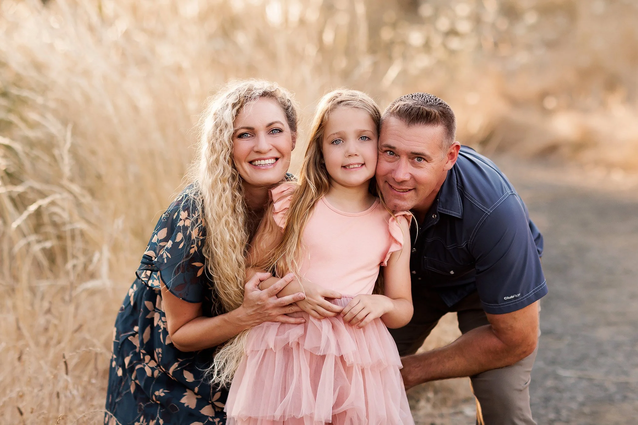 Parents kneeling with younger daughter during outdoor family photography session in warm golden light.