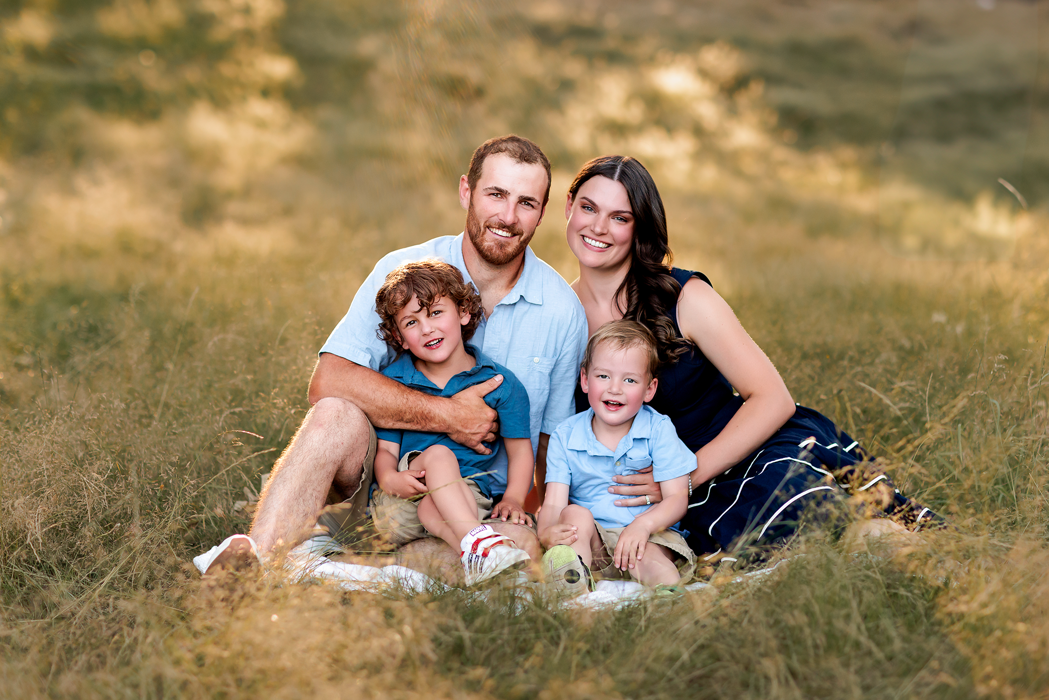 Family of four sitting together on a blanket in tall grass at golden hour, smiling at the camera