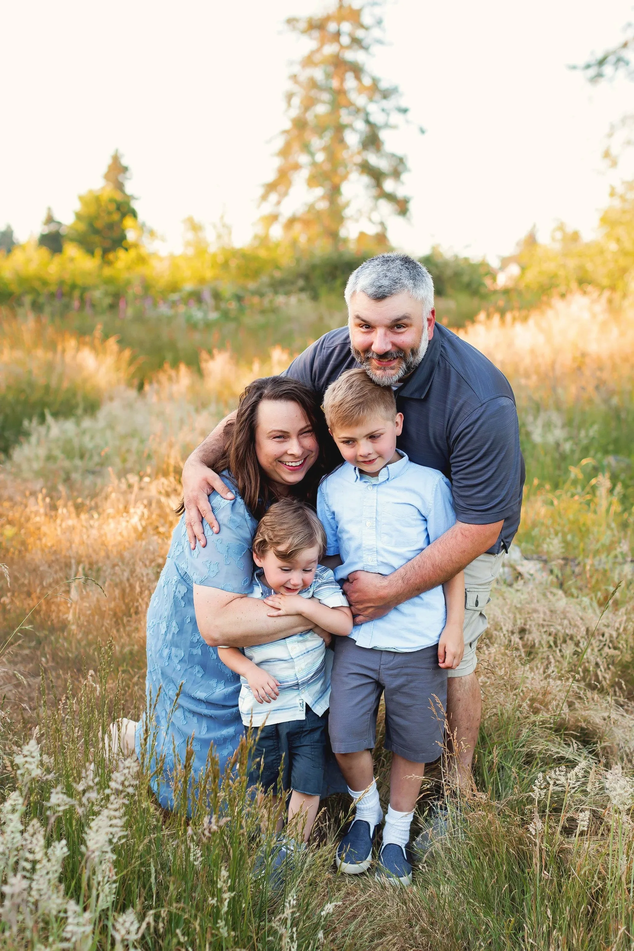 Family of four in golden field with mom kneeling and dad bending over hugging children close.