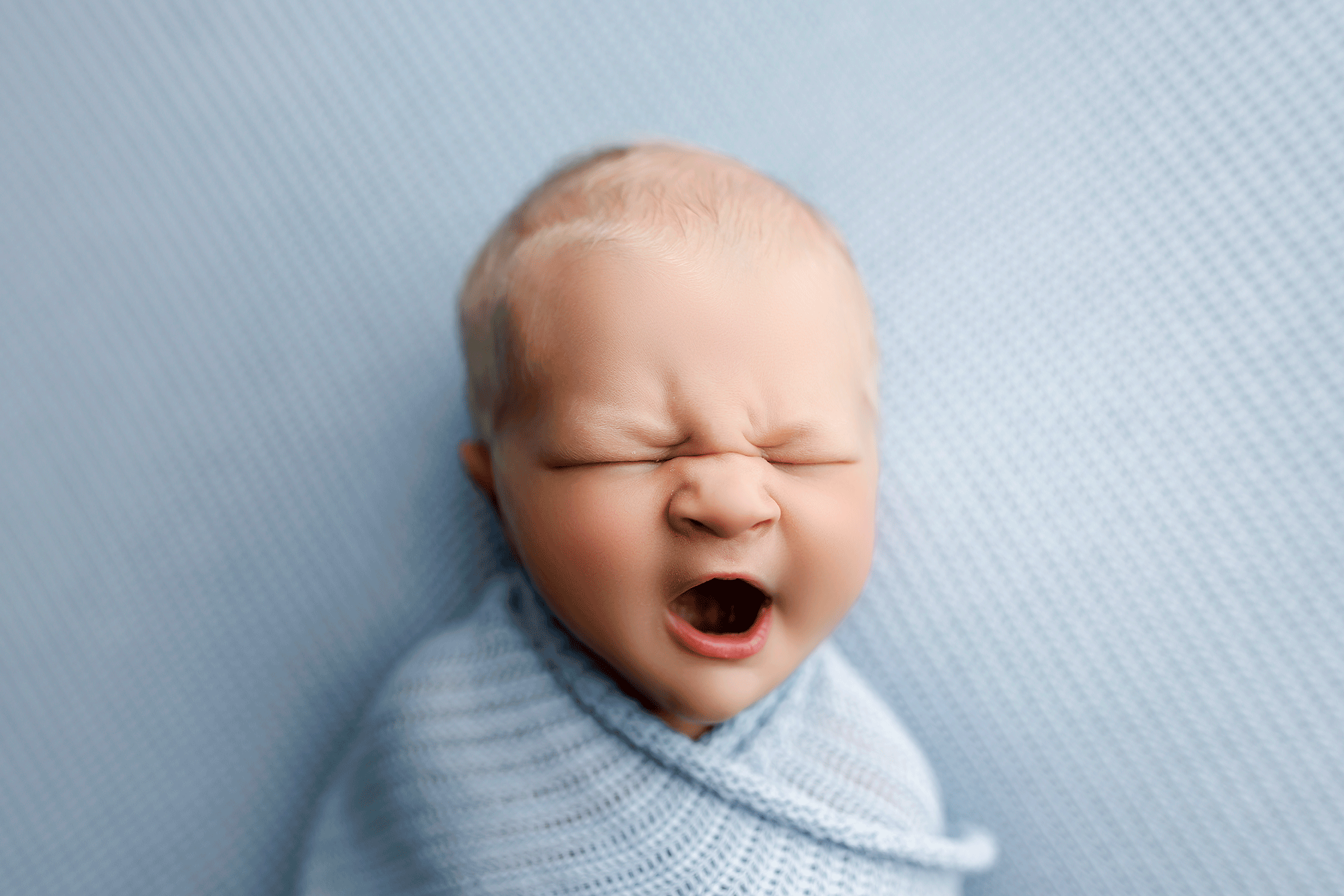 Close-up portrait of a yawning newborn baby wrapped in a soft blue blanket.