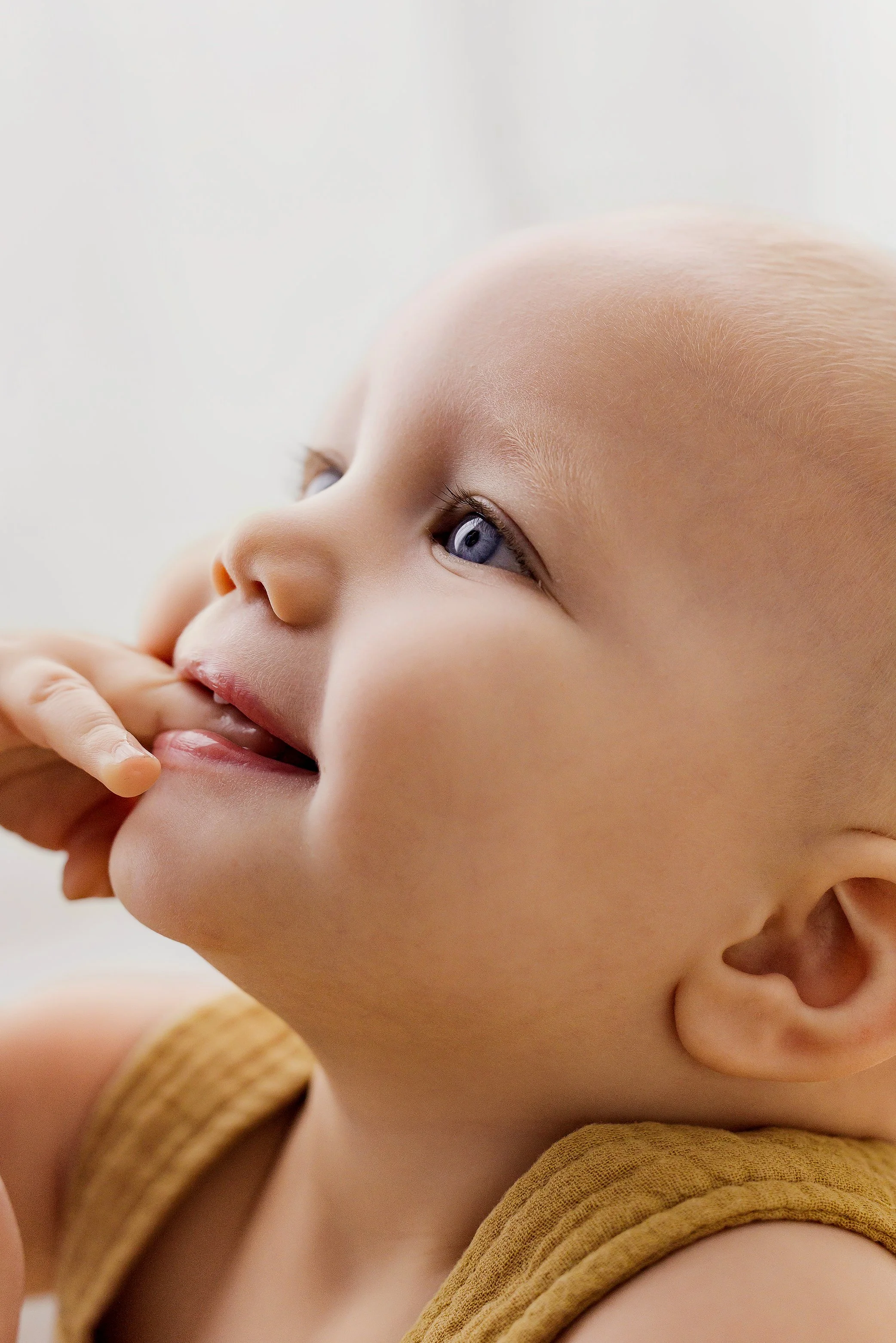 Close up of baby with blue eyes and finger in mouth during soft natural light baby photography session.