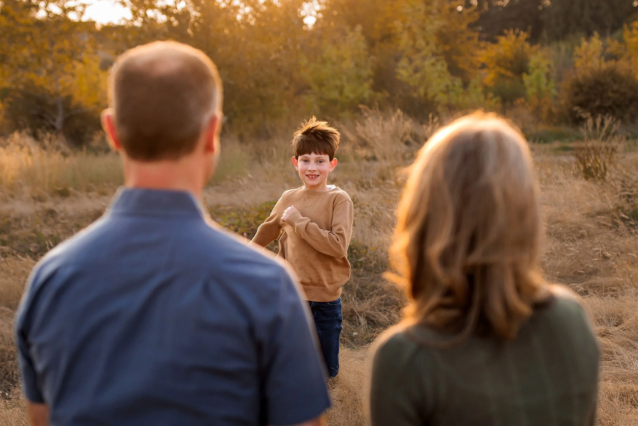 Young boy runs toward his parents who stand waiting in a sunlit field during golden hour.