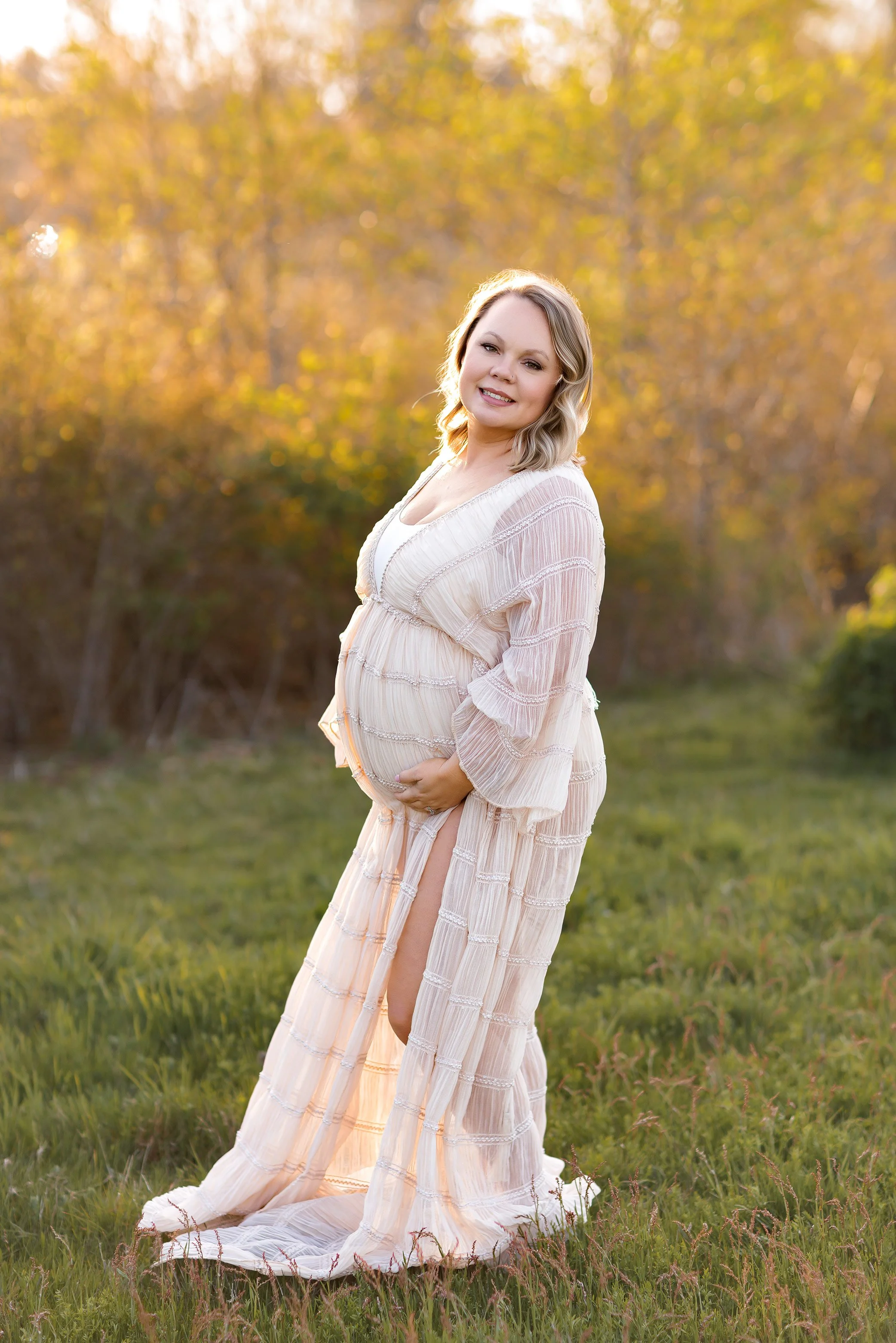 Expecting mother in flowing cream gown hugging her belly in golden field at sunset maternity session.