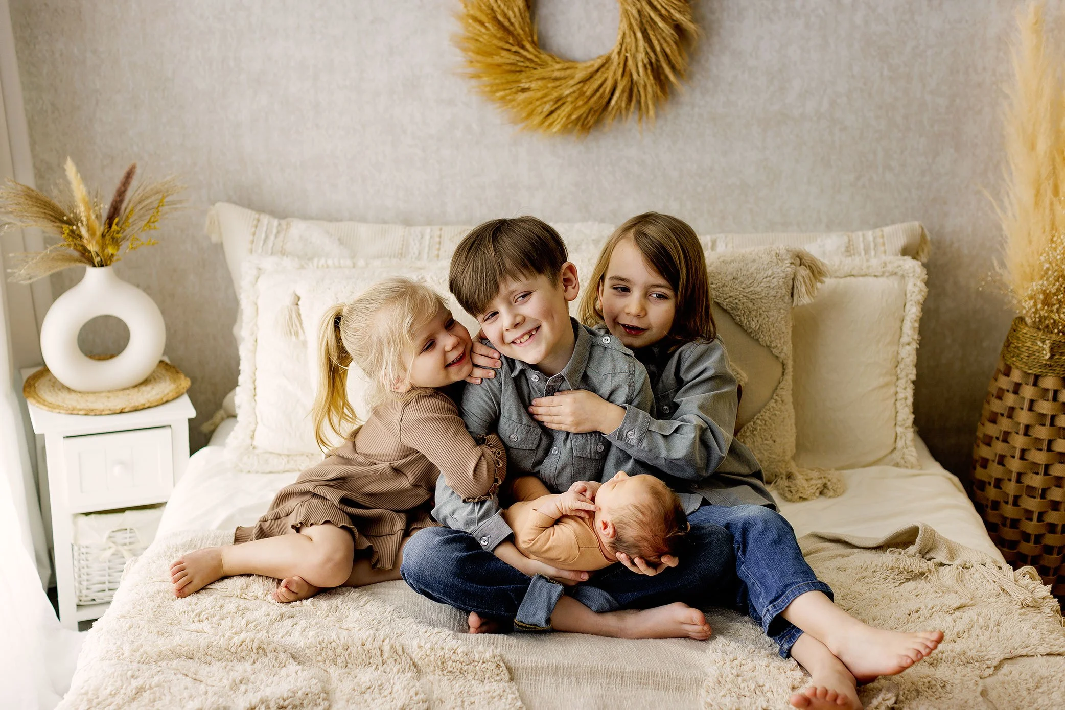 Three young siblings cuddling together on a bed while holding their newborn baby during a Salem, Oregon newborn photography session, capturing sibling connection and family love.