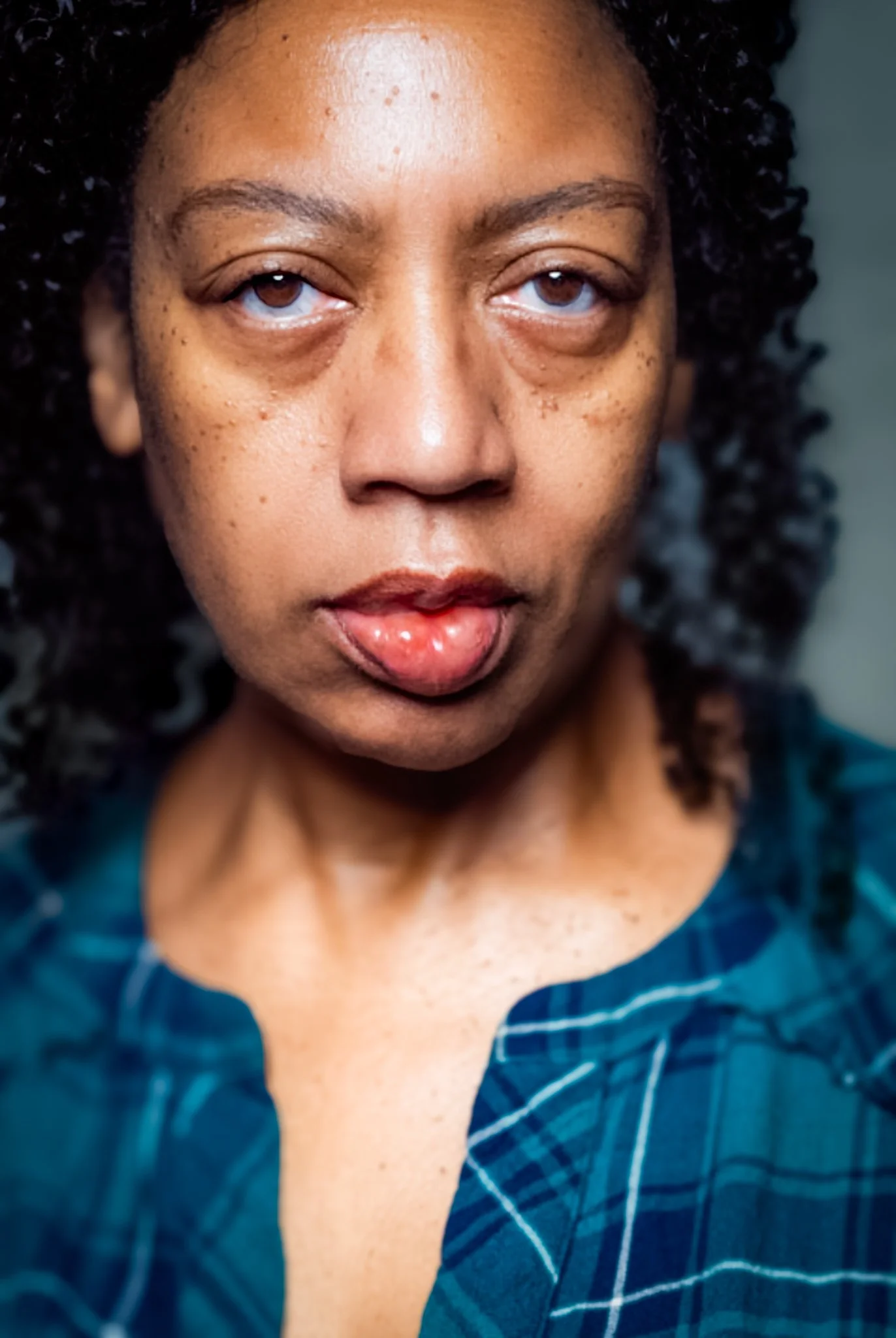 A close-up portrait of a woman with curly hair, light brown skin, and blue eyes, sticking out her tongue slightly, wearing a blue plaid shirt.