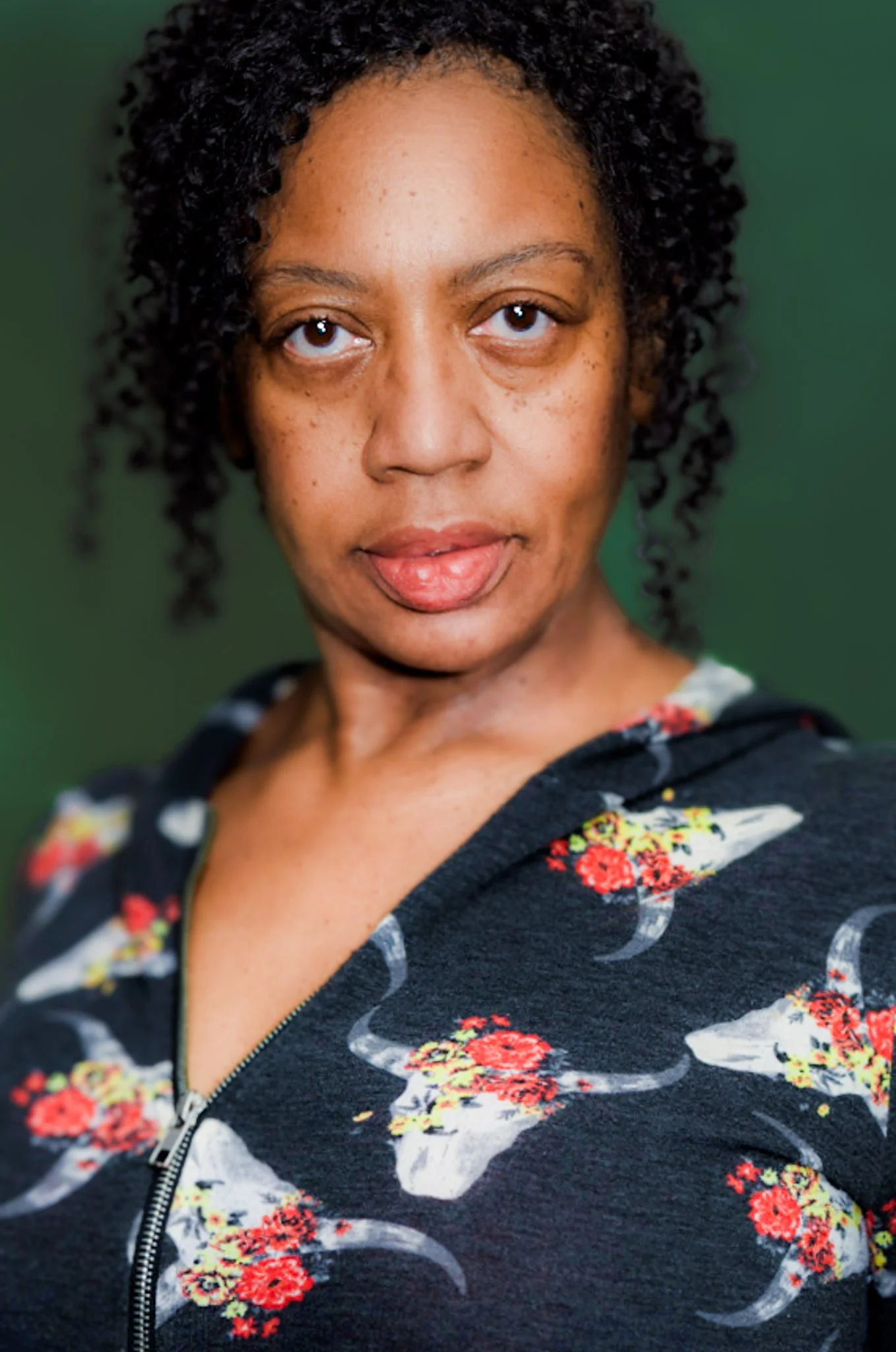 A woman with curly hair and brown skin looking directly at the camera against a green background, wearing a black zip-up hoodie with a fish skull and floral pattern.