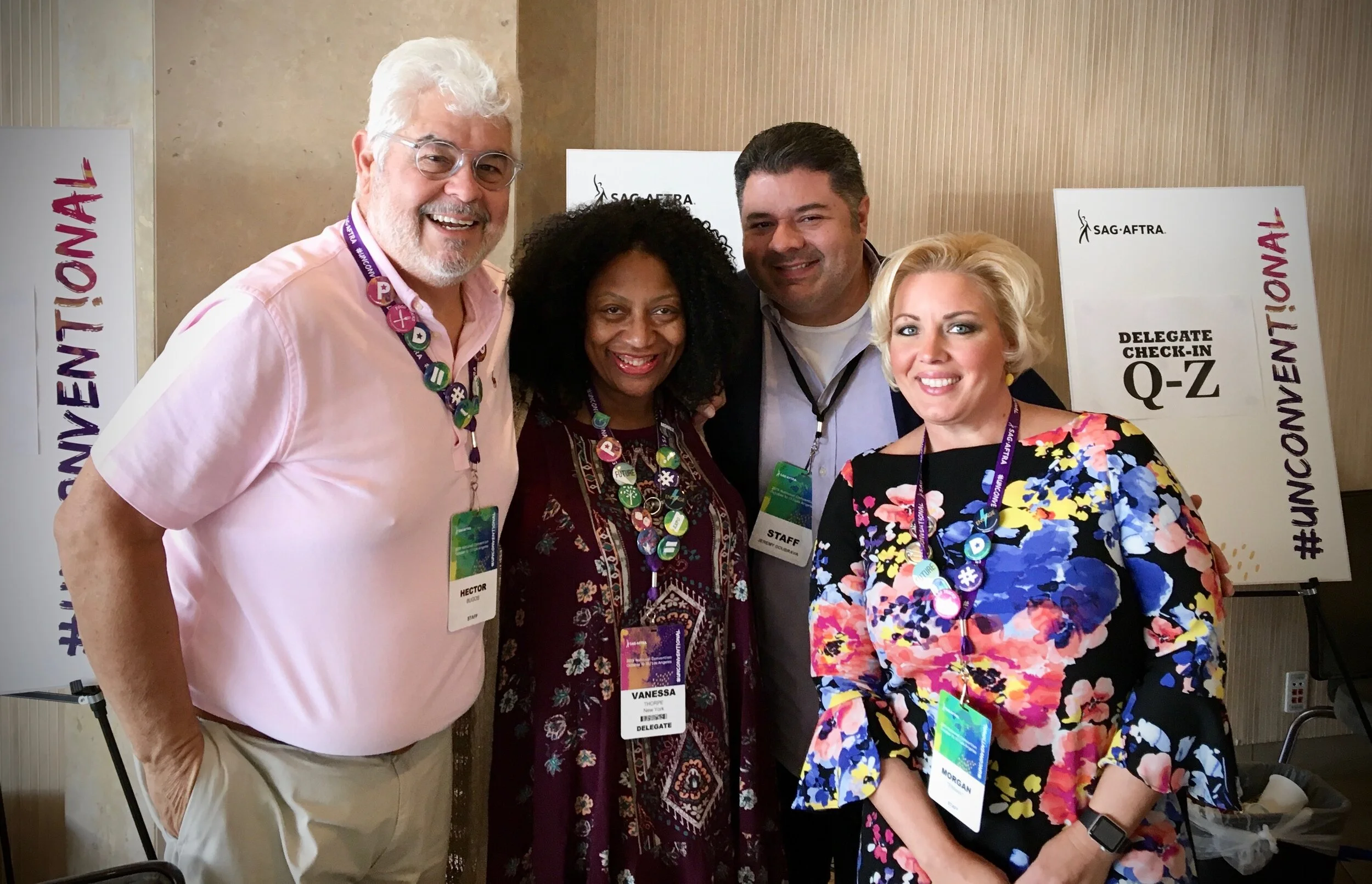 Four people at a SAG-AFTRA event, wearing badges and smiling, with signs in the background indicating delegate check-in.