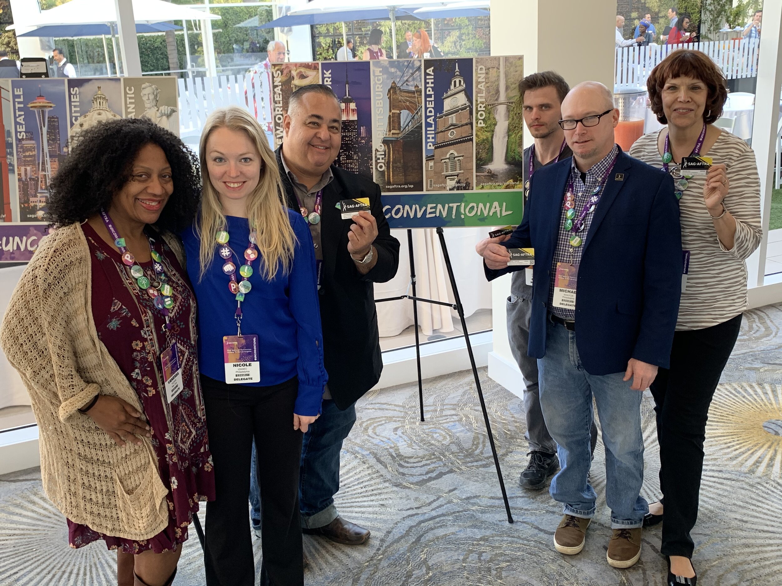 Group of six people at a convention, standing in front of a sign with city names and landmarks. They are wearing name tags and badges, smiling at the camera.