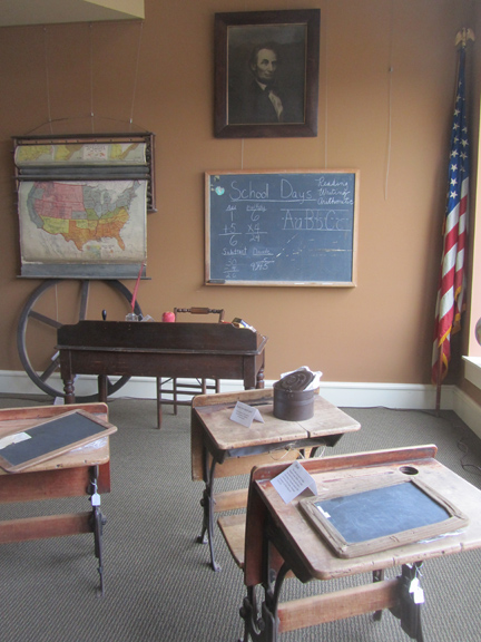 This museum gallery exhibit represents an early Lawrence County schoolroom. The wheel came from the bell tower at Central School. The maps were from the Springville School. At the beginning of the day, students recited the pledge of allegiance in front of an American flag such as the one shown in the corner.