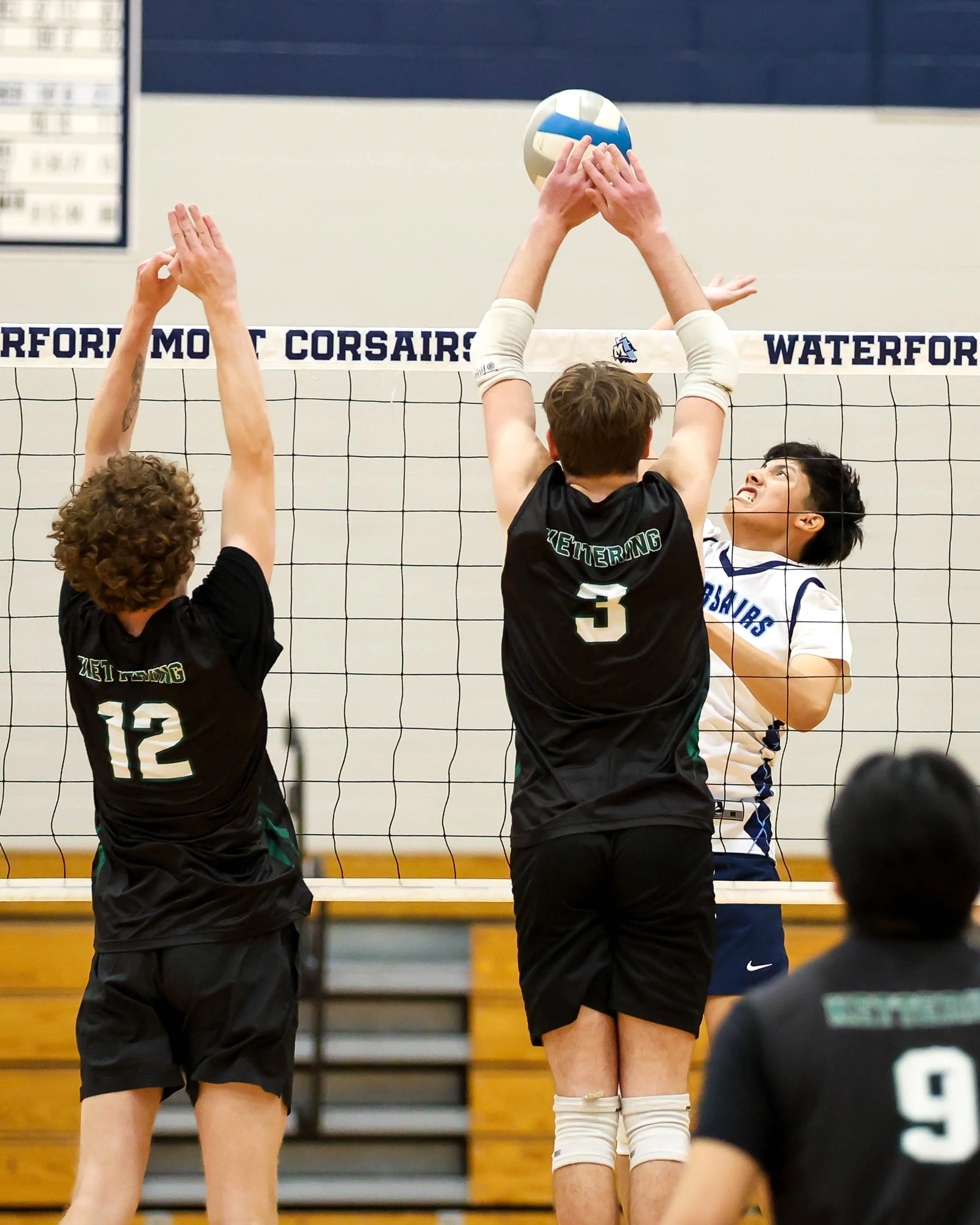 Mott #6 spikes the ball and Kettering #12 and #3 try to block it