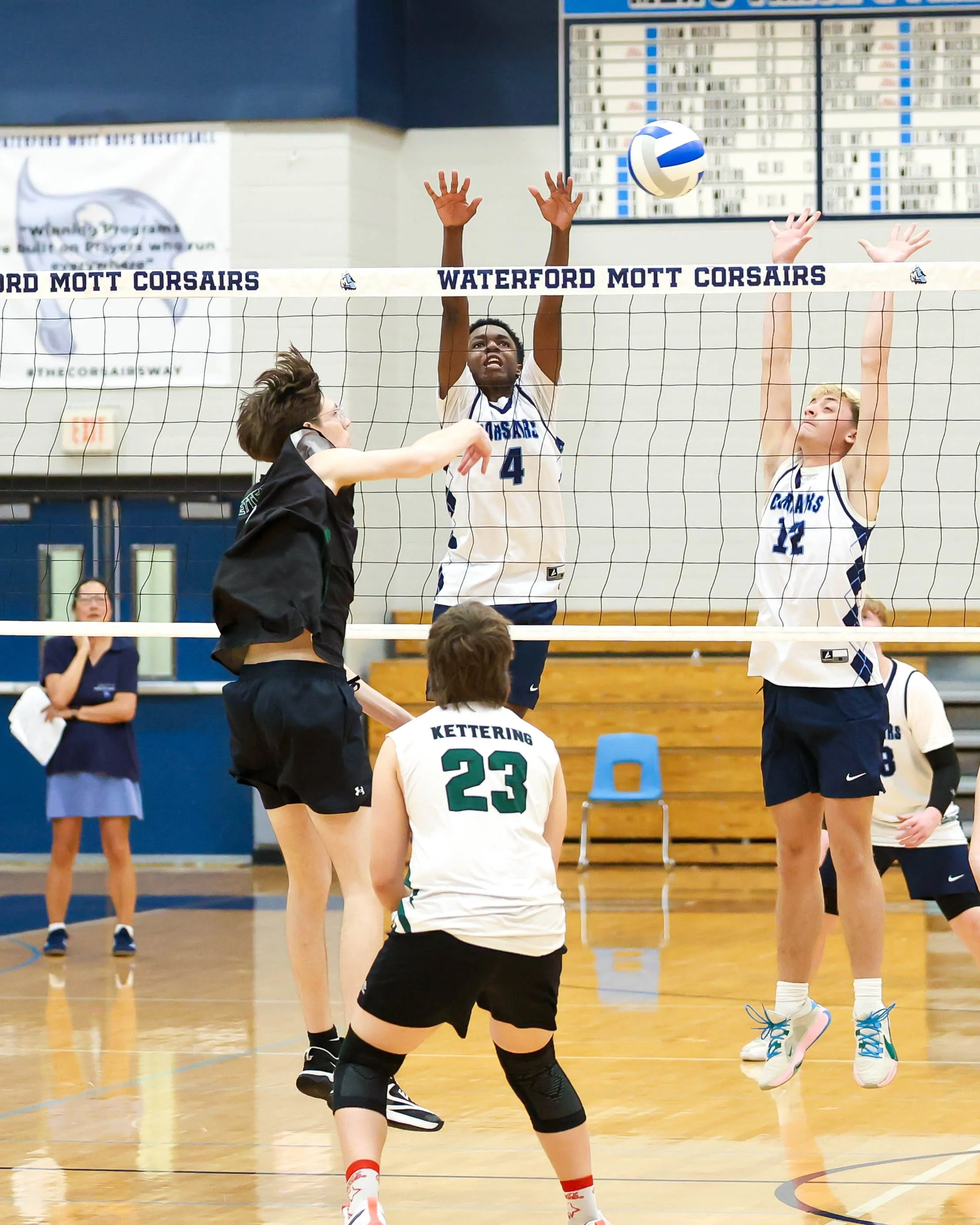Kettering #12 spikes the ball and Mott #4 and #18 try to block it
