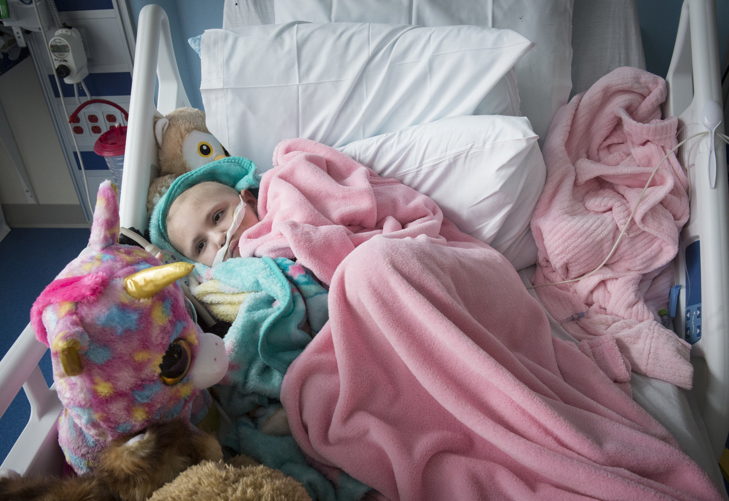  Naomi lays in her hospital bed, bundled up in blankets and surrounded by stuffed animals. 