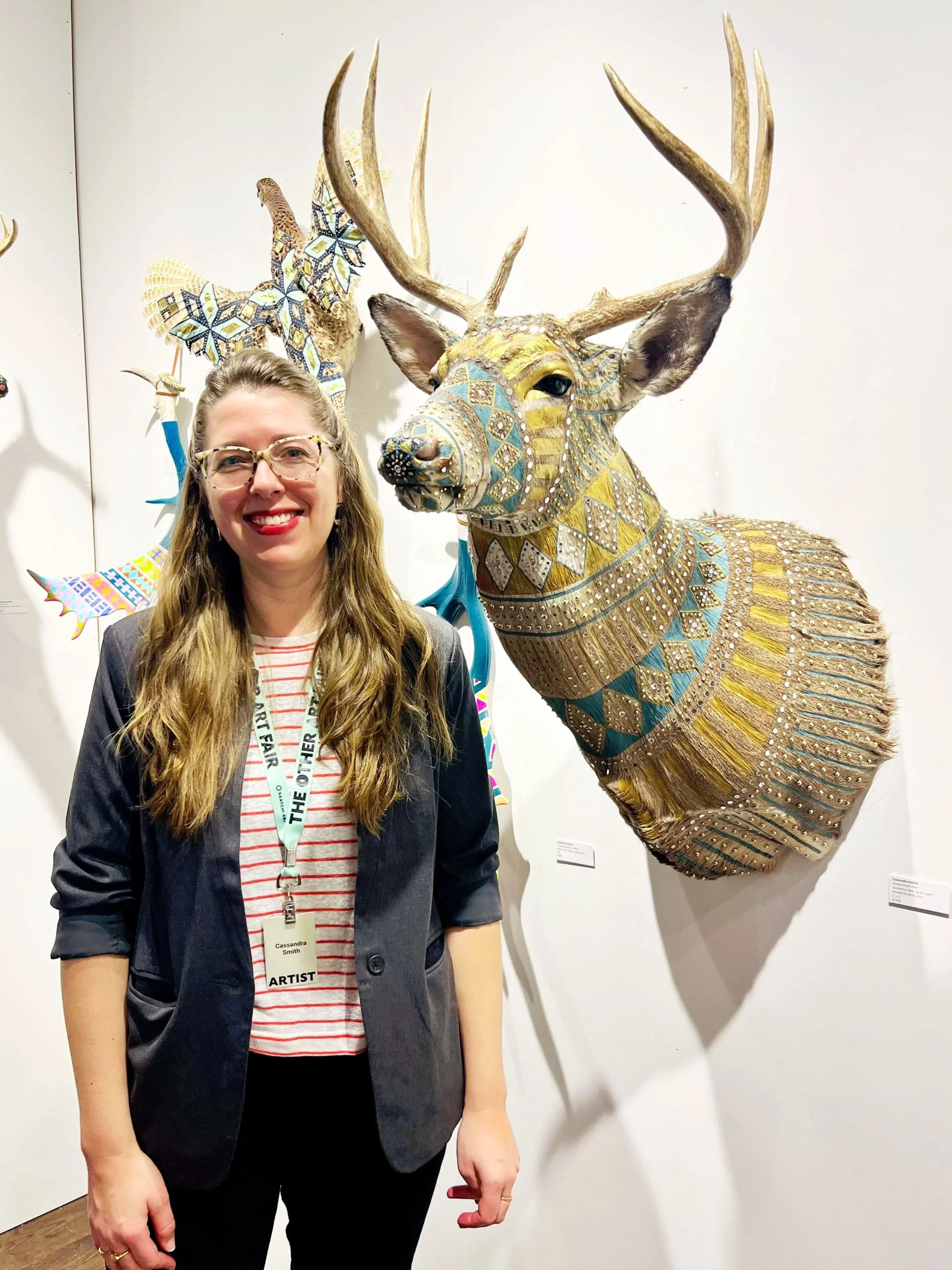 Artist Cassandra Smith standing in front of a large, decorated deer sculpture at an art exhibition.