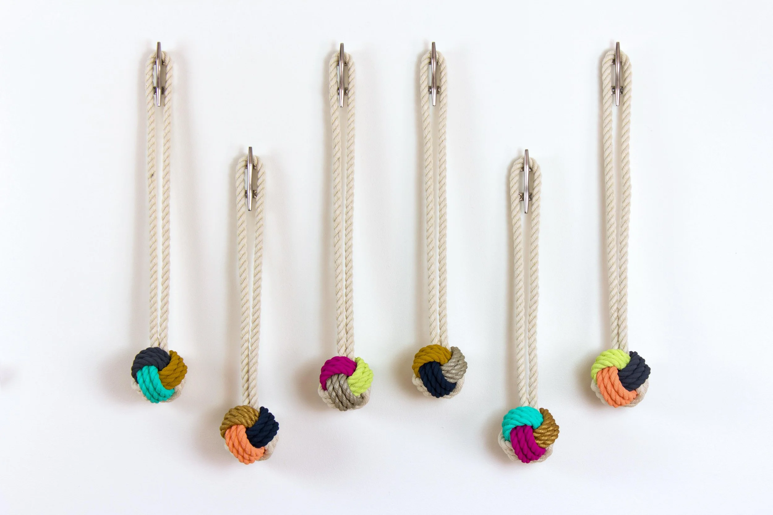 Six decorative ropes with hand-painted knots hanging on a white wall.