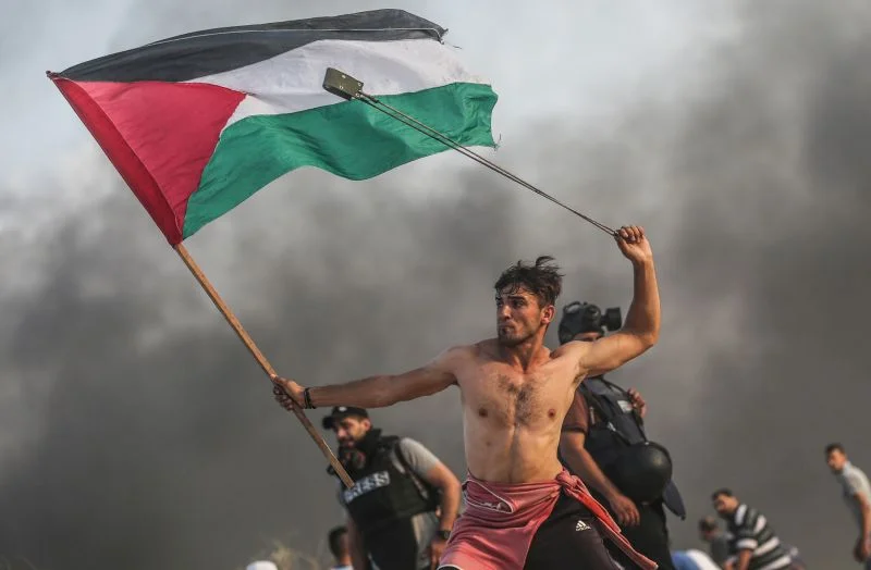 A shirtless young protester in Gaza gripping a Palestinian flag with one hand and swinging a slingshot over his head with the other, on the northern border between Gaza Strip and Israel in the weekly protests, which are organized by Palestinian protesters to protest against the Israeli blockade of Gaza which is imposed by Israel for 12 years until now. Photograph by Mustafa Hassona