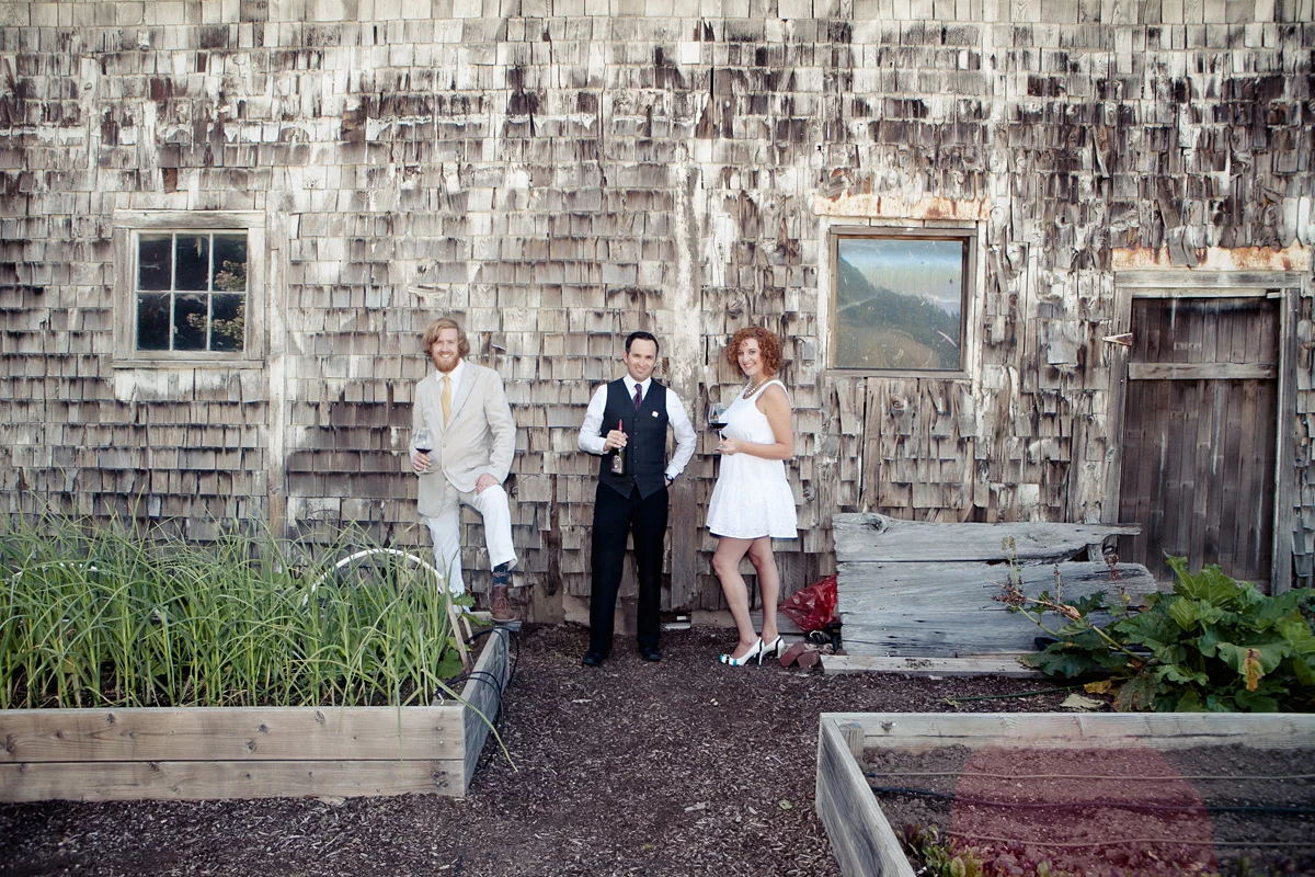 Three people standing outdoors in front of a weathered wooden building, holding wine glasses. There are garden beds with plants in the foreground and two windows on the building, with one person leaning against the wall.