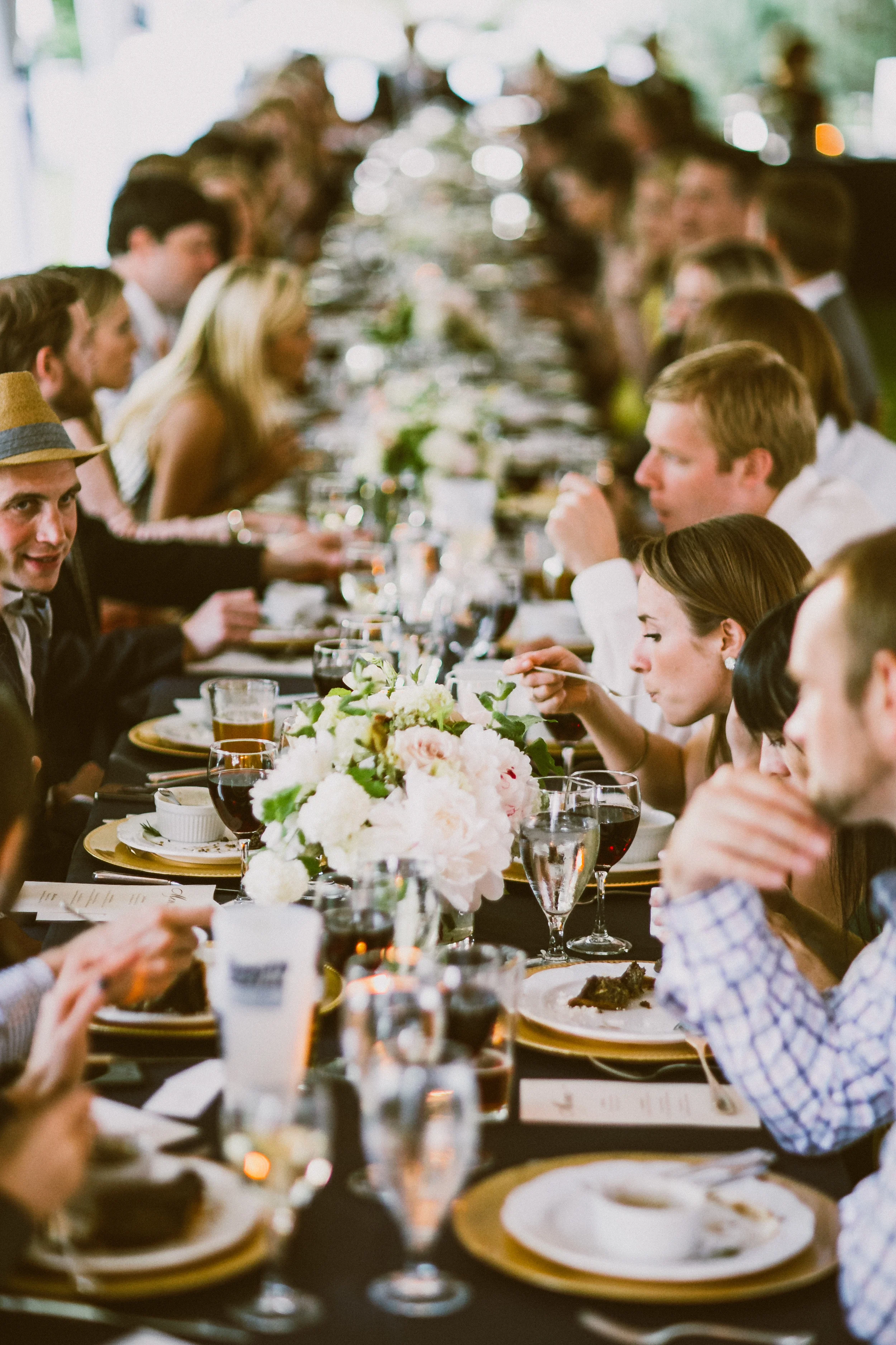 People sitting at a long banquet table decorated with flowers, enjoying a meal and socializing.