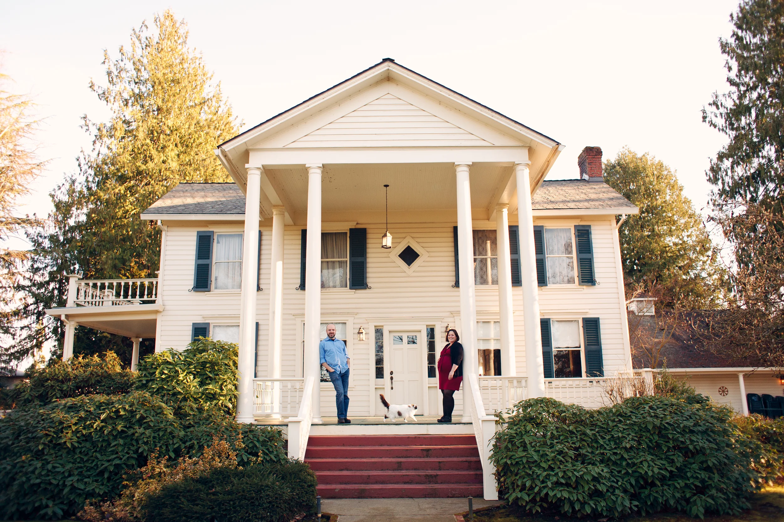 A colonial style house with a large front porch, white columns, and stairs leading up to the entrance. Two people and a small dog are standing on the porch, with lush bushes and trees in the background.