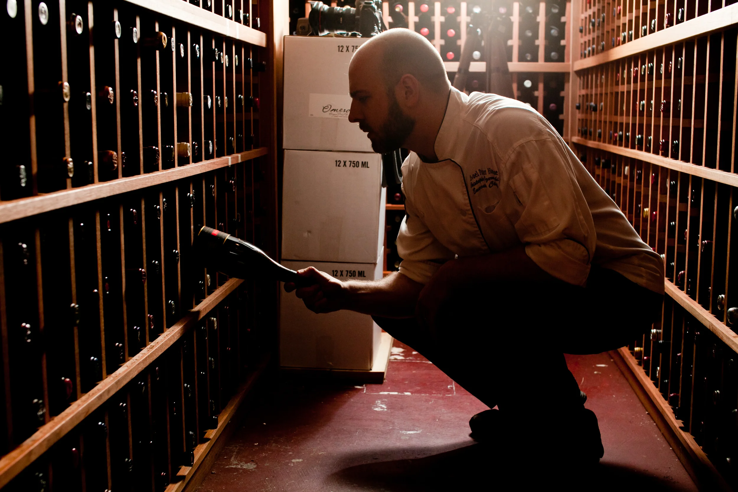 A man in a chef's coat squatting in a wine cellar, examining wine bottles on wooden shelves with a flashlight.