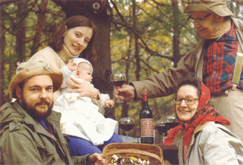 A family having a picnic outdoors in a forest with a woman holding a baby, a man with a beard wearing a hat, an elderly woman with glasses, and a man pouring wine.