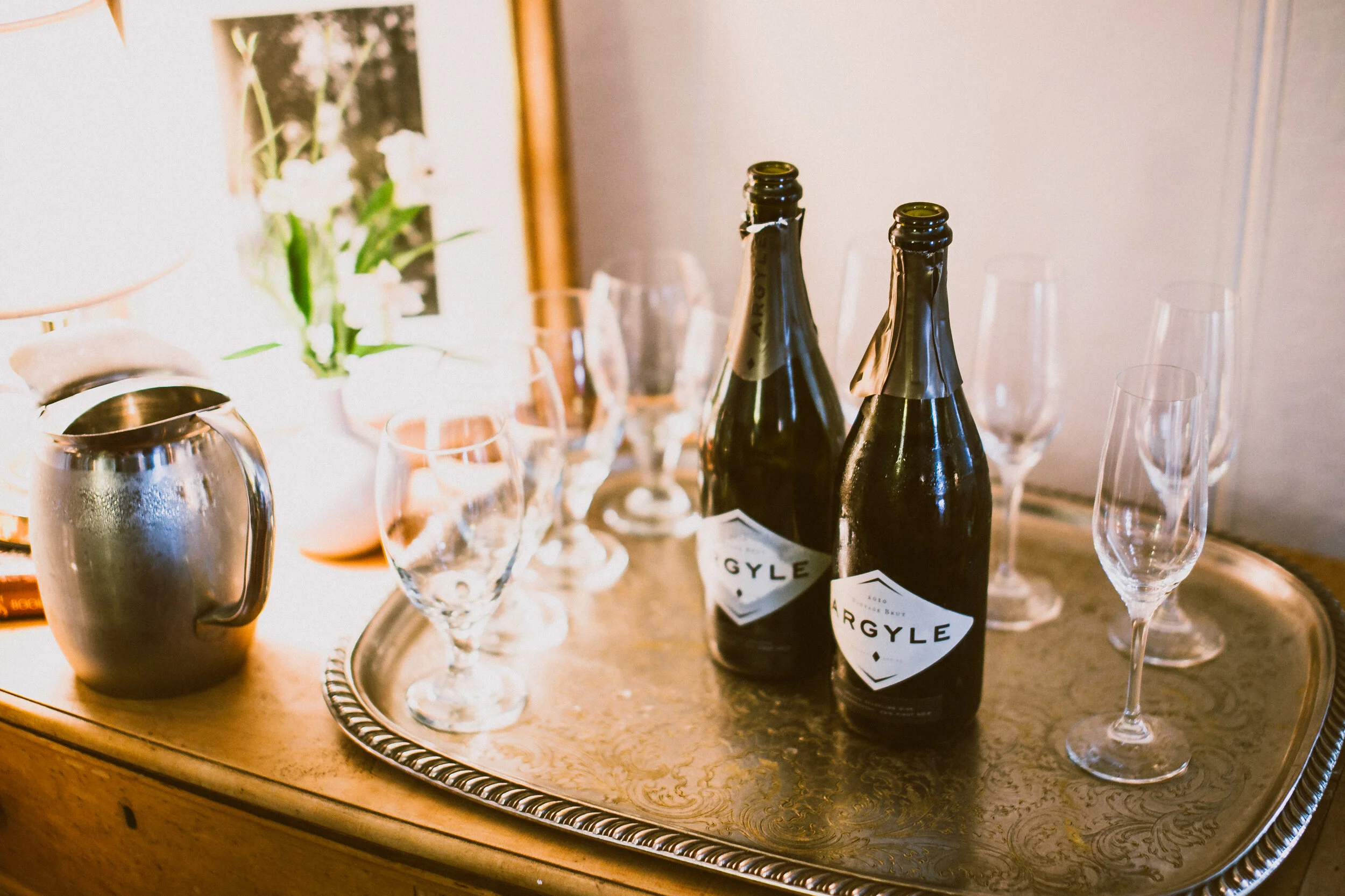 Two bottles of Argyle sparkling wine, several empty champagne glasses, a silver ice bucket, and a tray with a decorative edge sit on a wooden bar or side table.