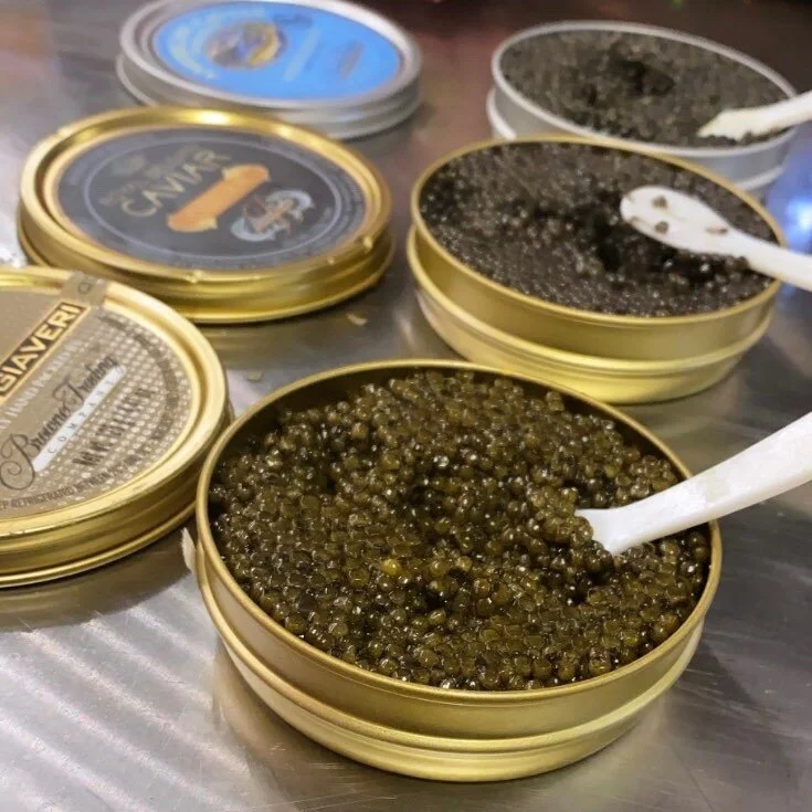 Several small metal tins filled with fish roe, including black caviar and greenish capelin roe, with plastic spoons for serving.