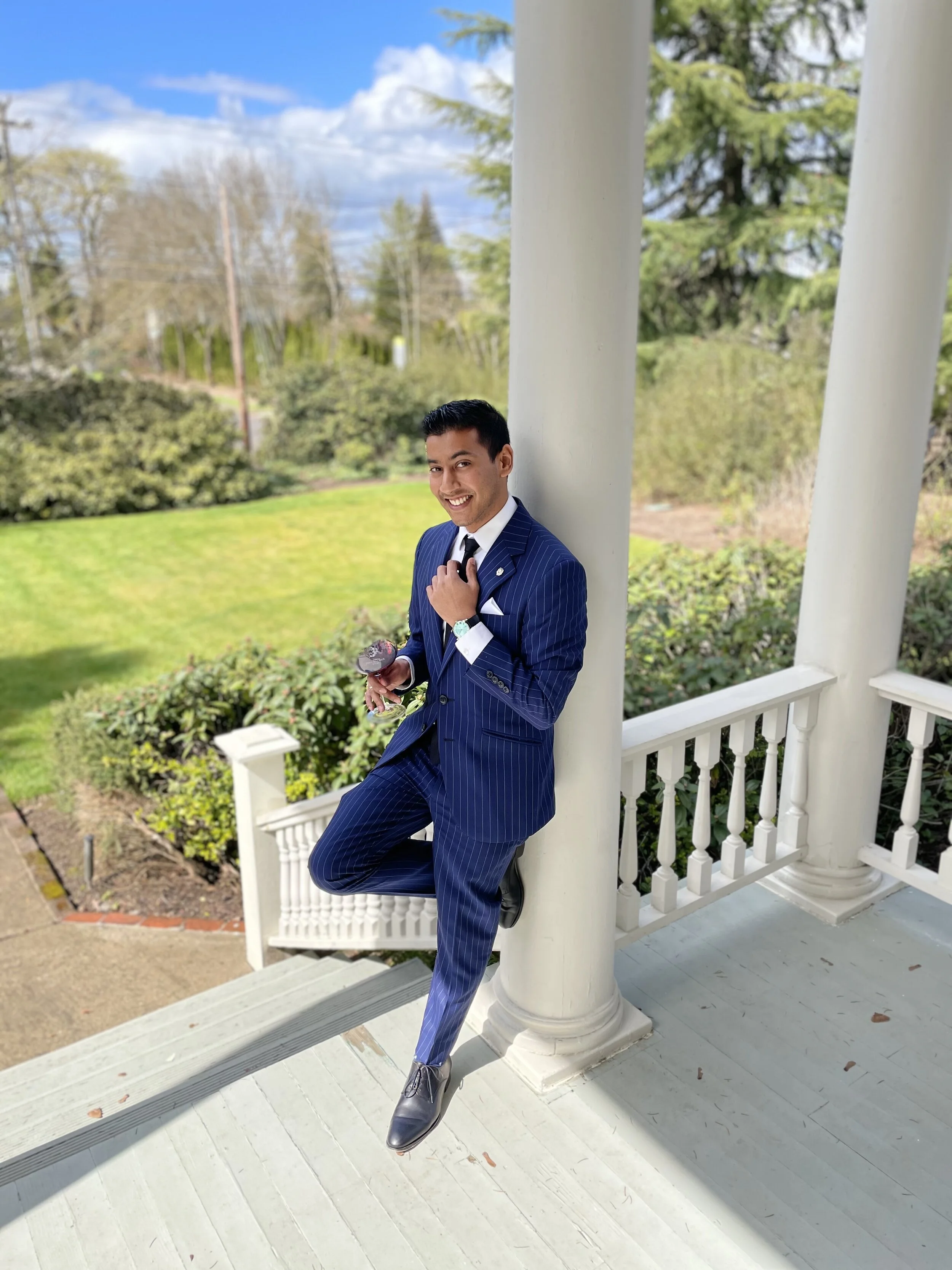 A man in a blue pinstripe suit standing on a porch, holding a glass of wine, smiling, with greenery and trees in the background.
