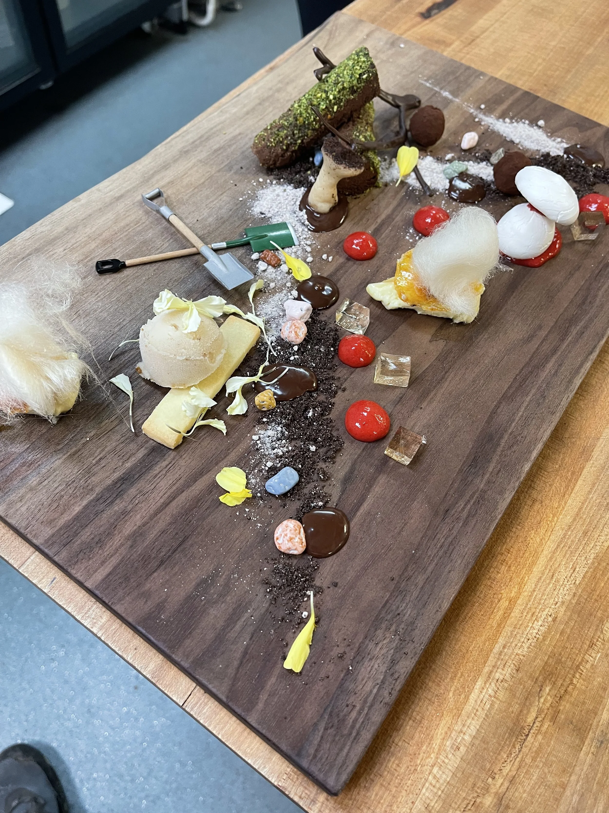 An art piece on a wooden board made with various food items, candies, and small tools, featuring chocolate, marshmallows, and garnishes in a creative arrangement.