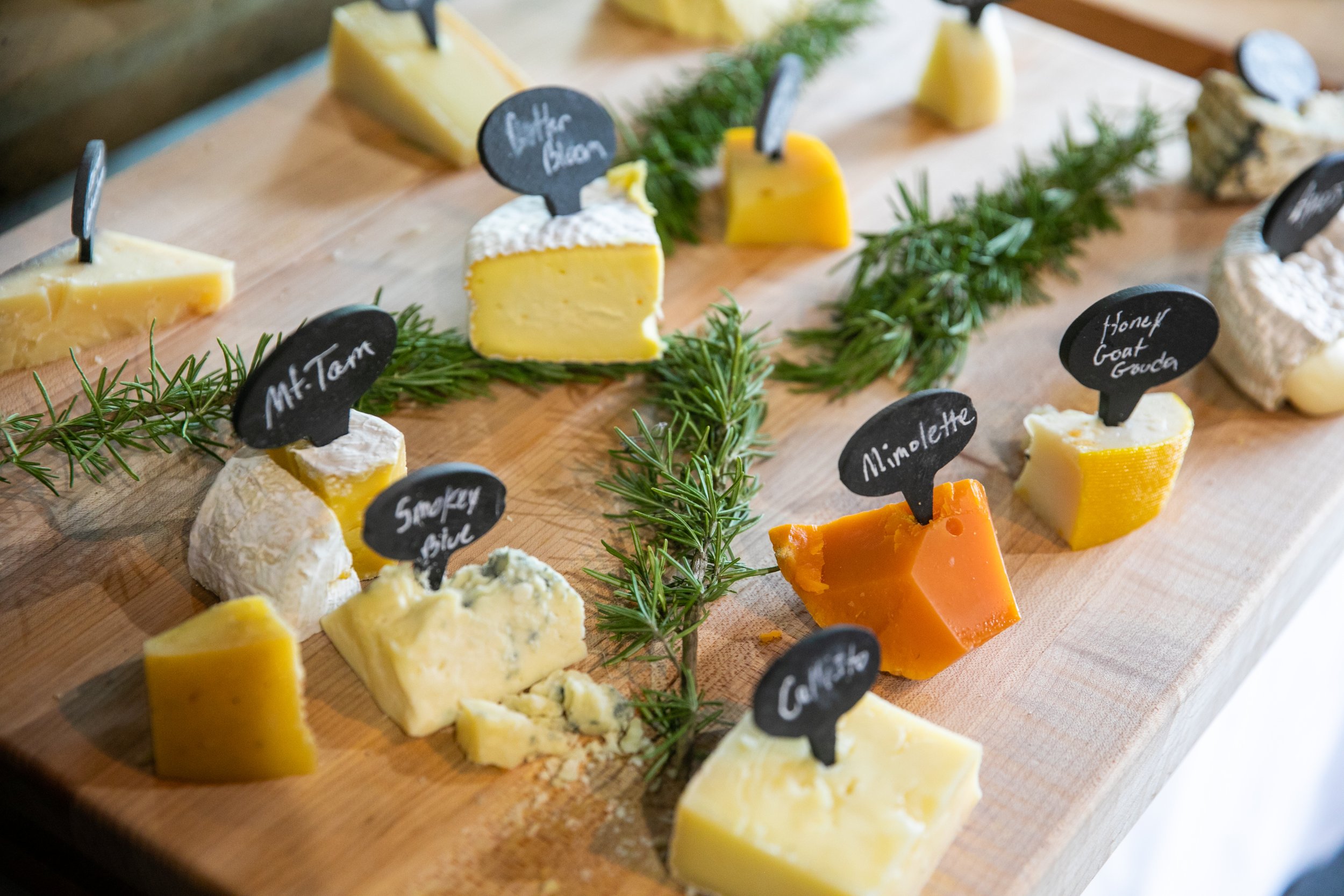 Assorted cheeses on a wooden cheese board with small black labels indicating names like "Mt. Tam," "Smokey Side," "Gouda," "Mimolette," and others, garnished with sprigs of rosemary.