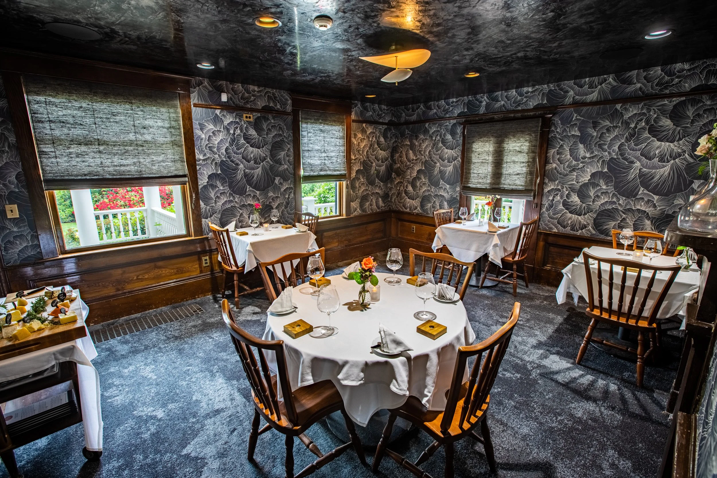 Interior of a cozy restaurant dining room with round tables covered in white tablecloths, each set with glassware, napkins, and floral centerpieces. The walls are decorated with dark floral wallpaper, and four windows with blinds let in natural light