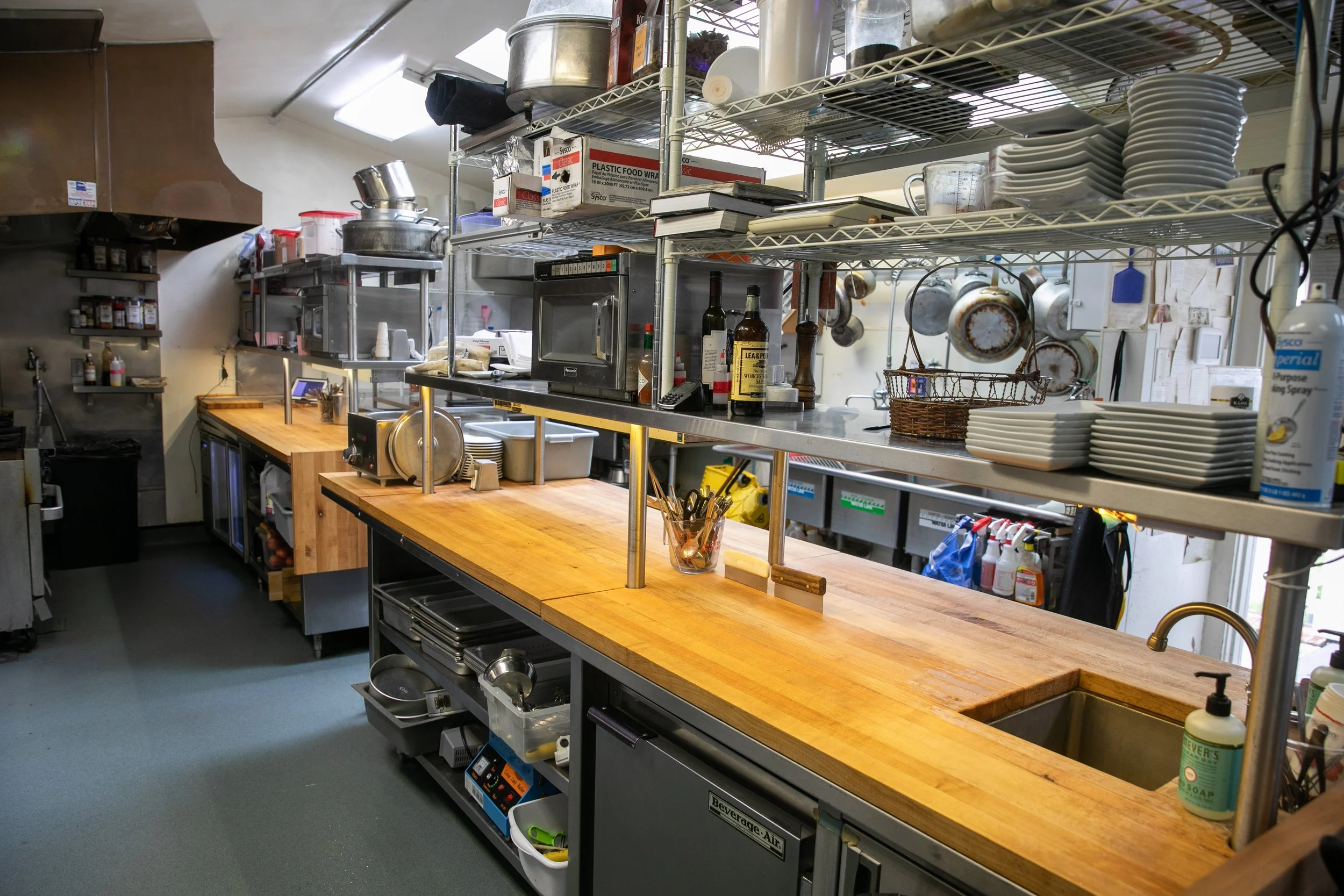 A commercial kitchen with a long wooden counter, open shelving with plates, bowls, utensils, and kitchen tools, and various appliances and supplies such as a microwave, bottles of wine, and cleaning products.