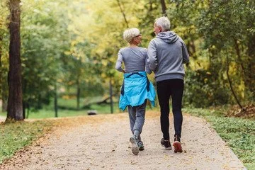 Two elderly women walking together along a dirt path in a park, surrounded by trees with green and yellow leaves.
