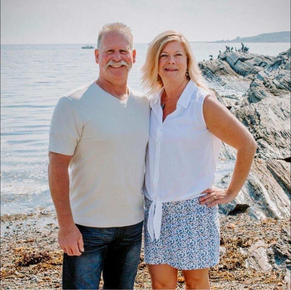 A smiling older man and woman standing on a rocky beach near the water, with a boat and distant shoreline in the background.