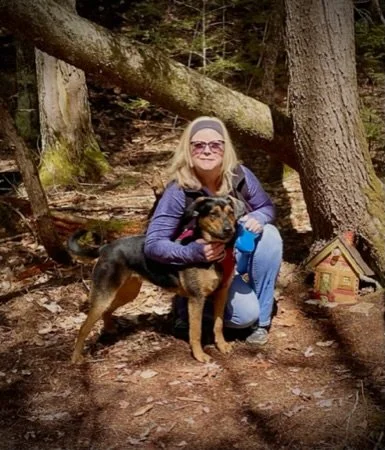 A woman with blonde hair, wearing sunglasses, a purple jacket, and jeans, kneels next to a large dog in a wooded area with trees and a small wooden house figure nearby.