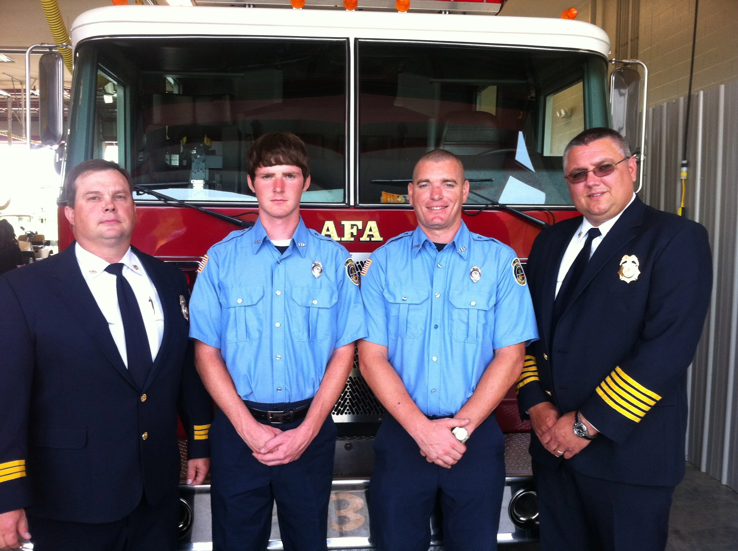 Four firefighters standing in front of a fire truck inside a fire station. The two on the left are in light blue uniforms, and the two on the right are in dark navy uniforms.