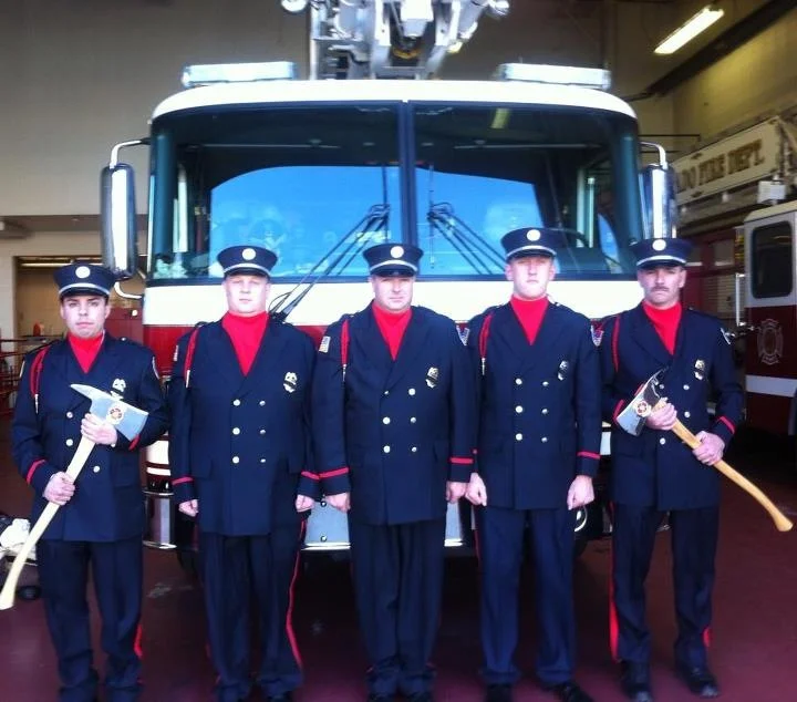 Five young firefighters in formal navy uniforms with red accents and hats, standing in front of a fire truck inside a fire station.
