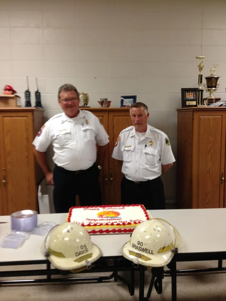 Two firefighters in uniform standing behind a table with a cake, helmets, and awards in a room with wooden cabinets and trophies.