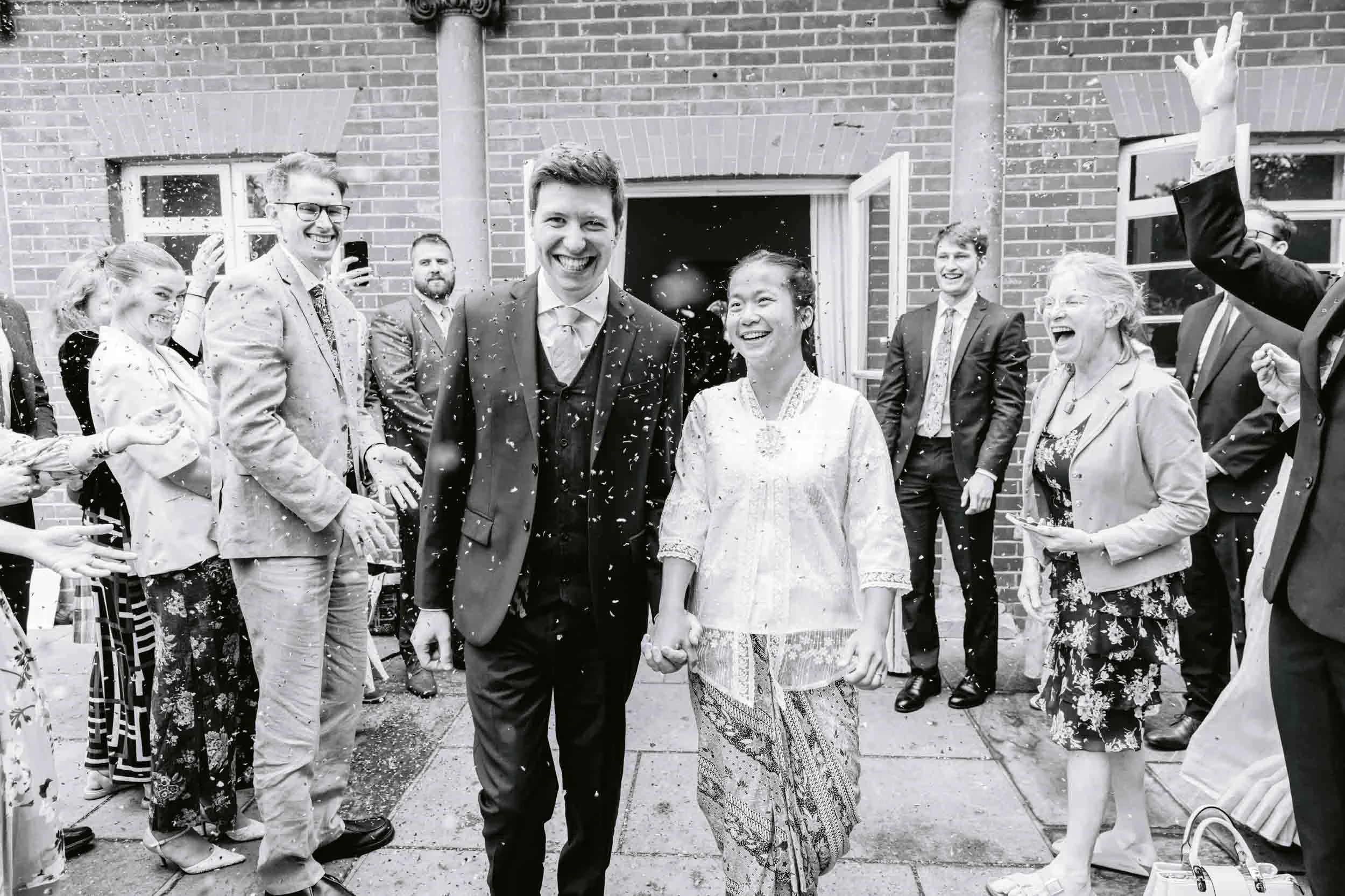 A wedding celebration with a bride and groom holding hands and smiling, surrounded by friends and family throwing confetti outside a brick building.
