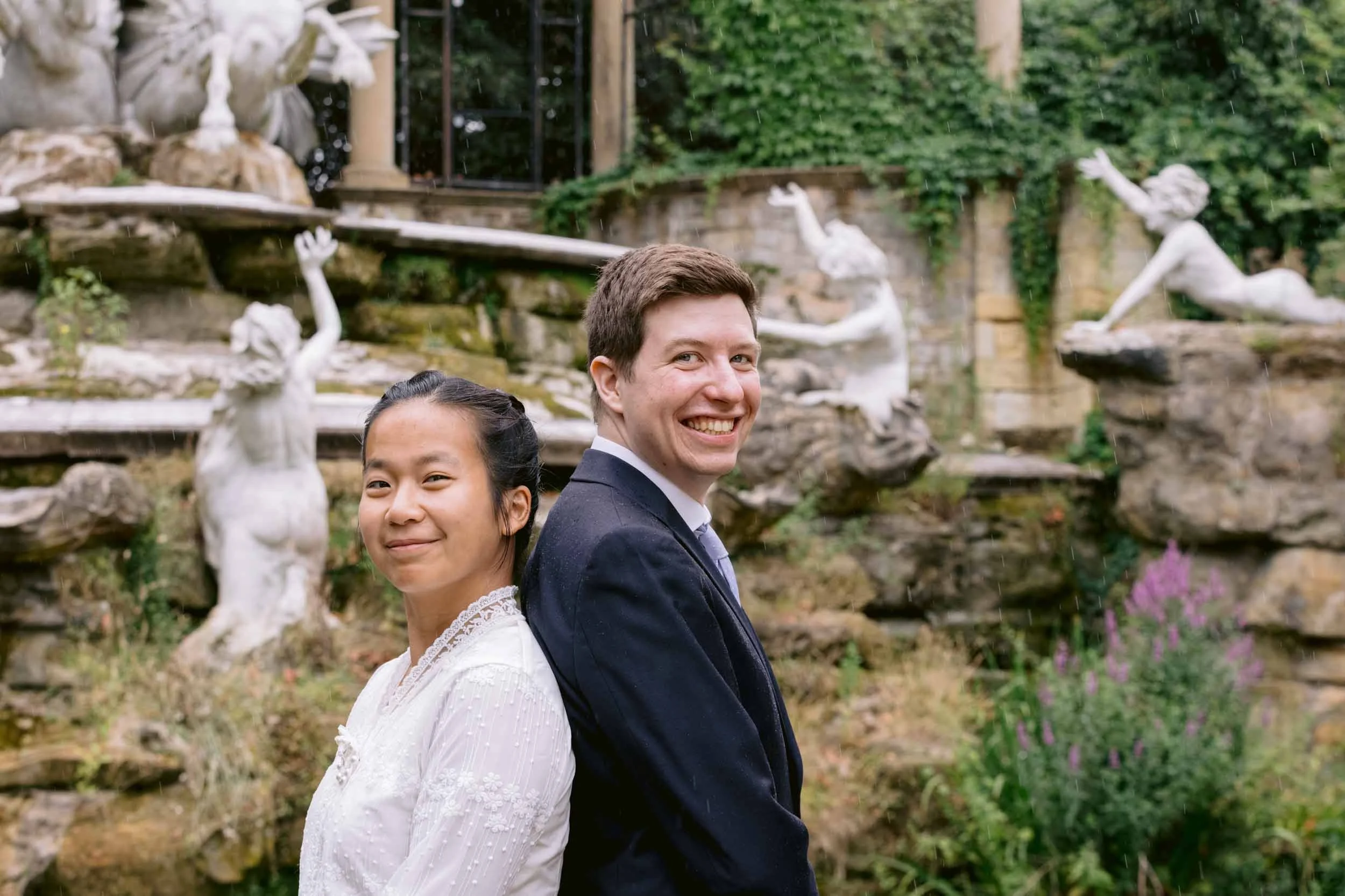 Two smiling people, a woman and a man, standing back to back outdoors during light rain, with decorative white statues and lush green foliage in the background.