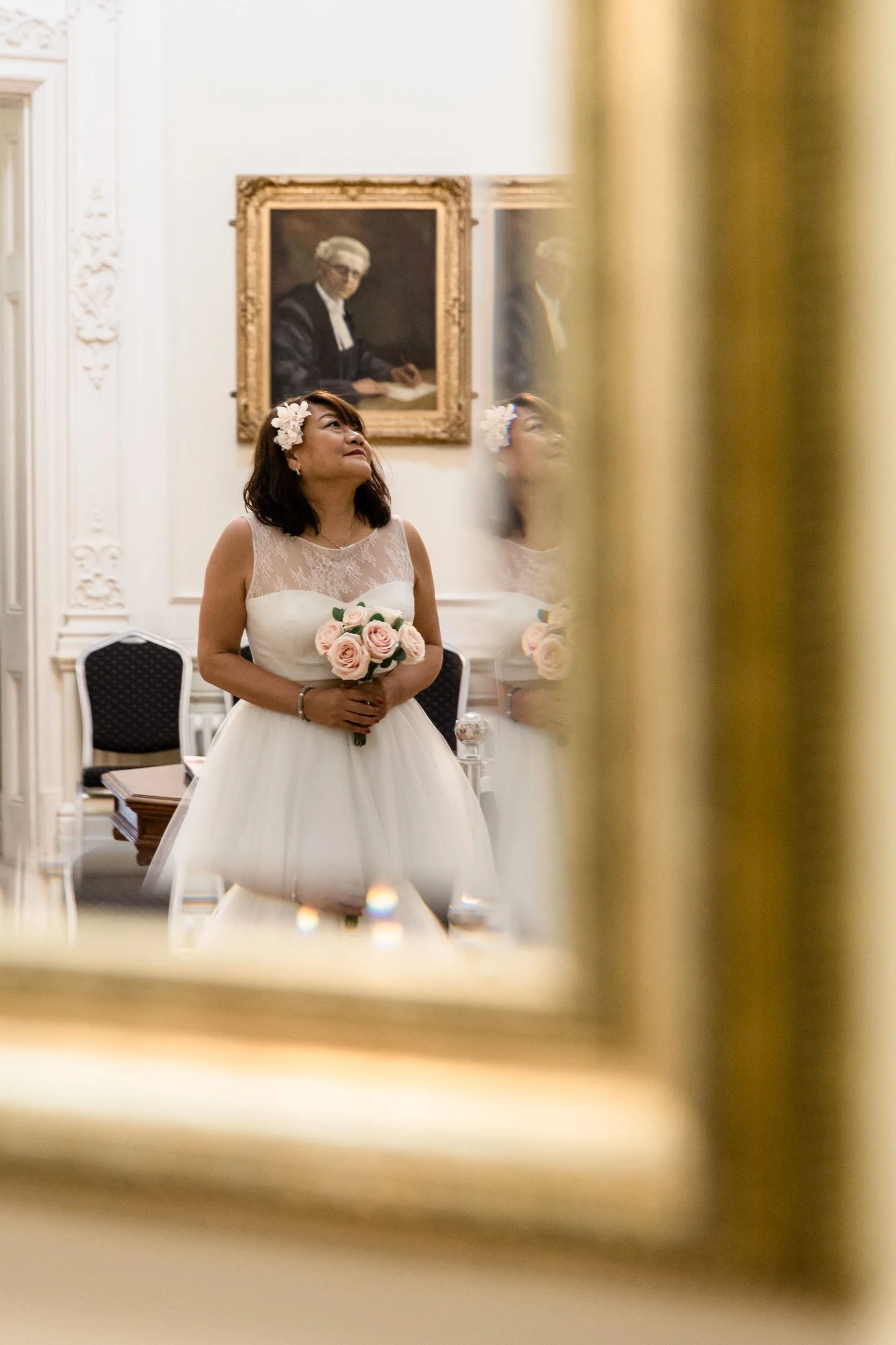 A woman in a wedding dress with flowers in her hair holding a bouquet of pink roses, looking up inside a room with ornate white walls and framed portraits.