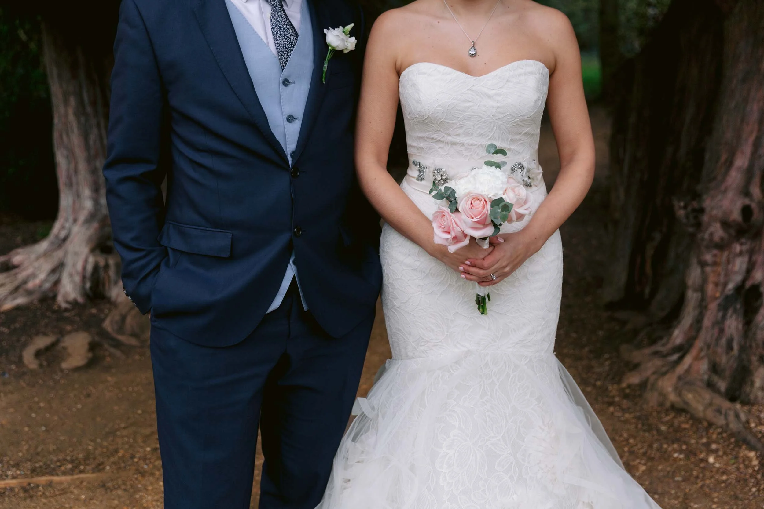 Close-up of a bride and groom standing outdoors, showing their torsos. The groom is wearing a navy blue suit with a light blue vest and a patterned tie. The bride is wearing a strapless white lace wedding dress with a sweetheart neckline, holding a bouquet of pink roses and greenery.