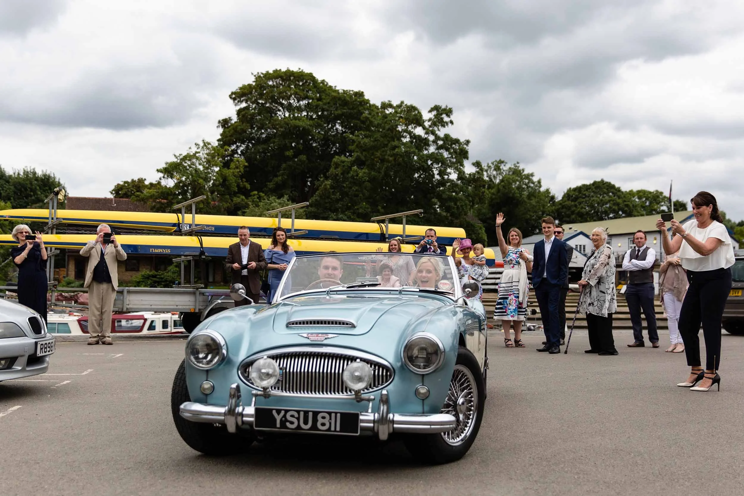 A light blue vintage sports car with a black and white license plate YSU 811 is driving towards the camera as people enjoy a gathering in the background. The crowd is taking photos of the car and waving, with overcast skies above.