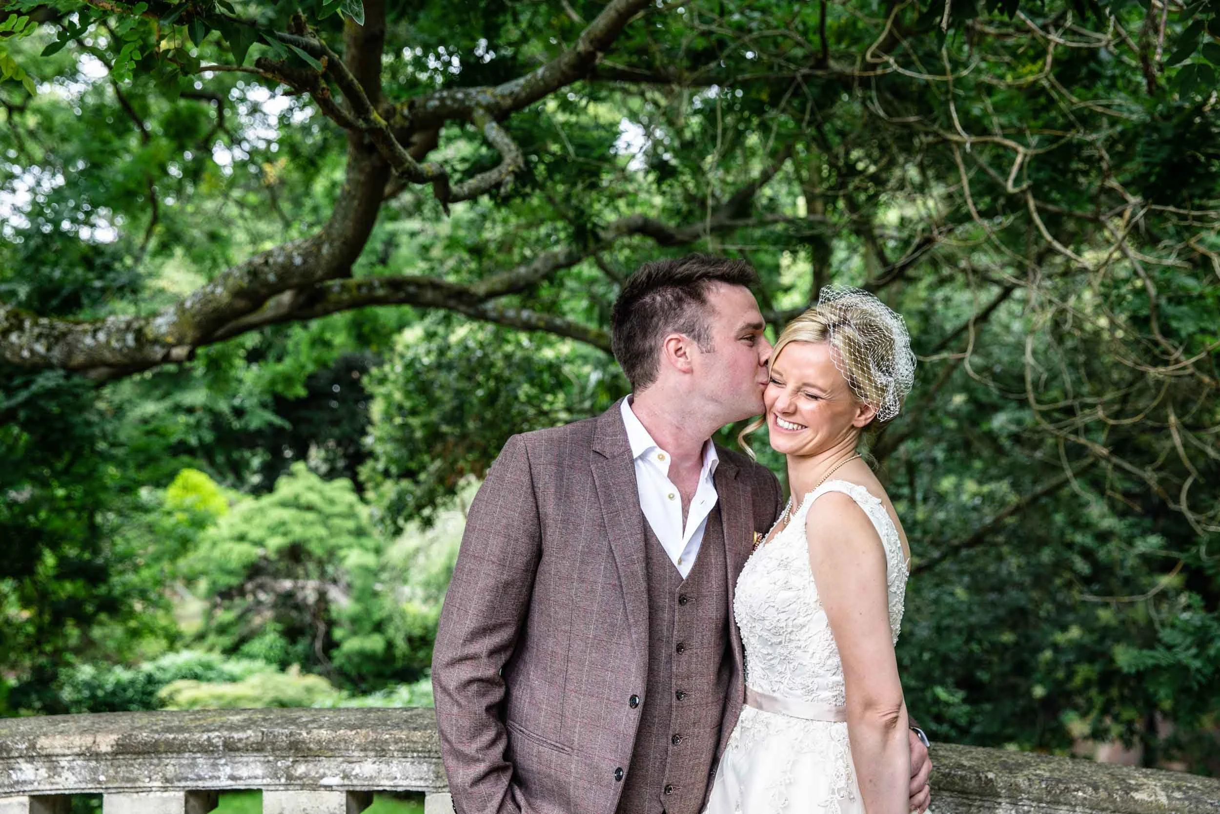 A newlywed couple standing outdoors in front of a large tree, with the groom kissing the bride on the cheek. The bride is smiling and wearing a wedding dress with a birdcage veil, while the groom is dressed in a brown plaid suit with a white shirt.