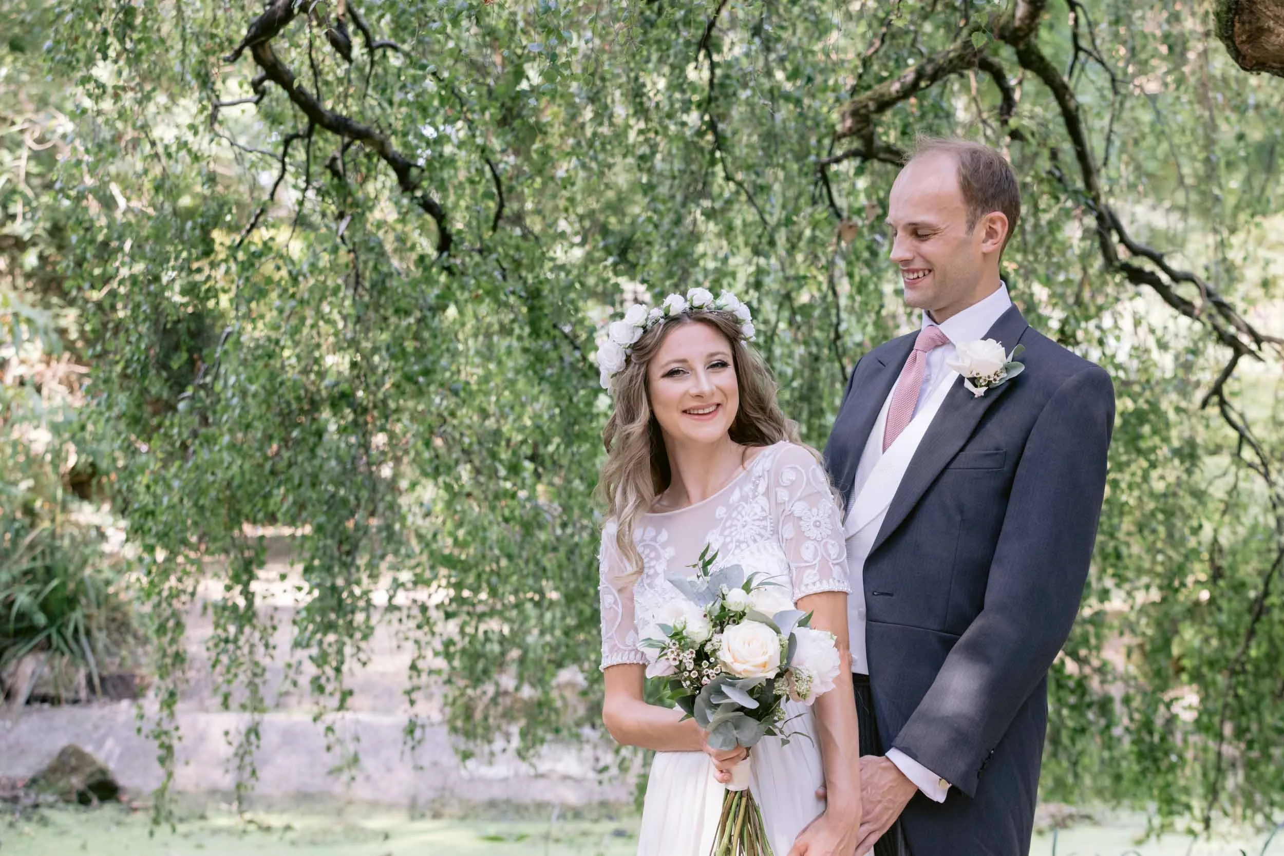 A bride and groom smiling outdoors, holding a bouquet of white roses, with green trees and foliage in the background.