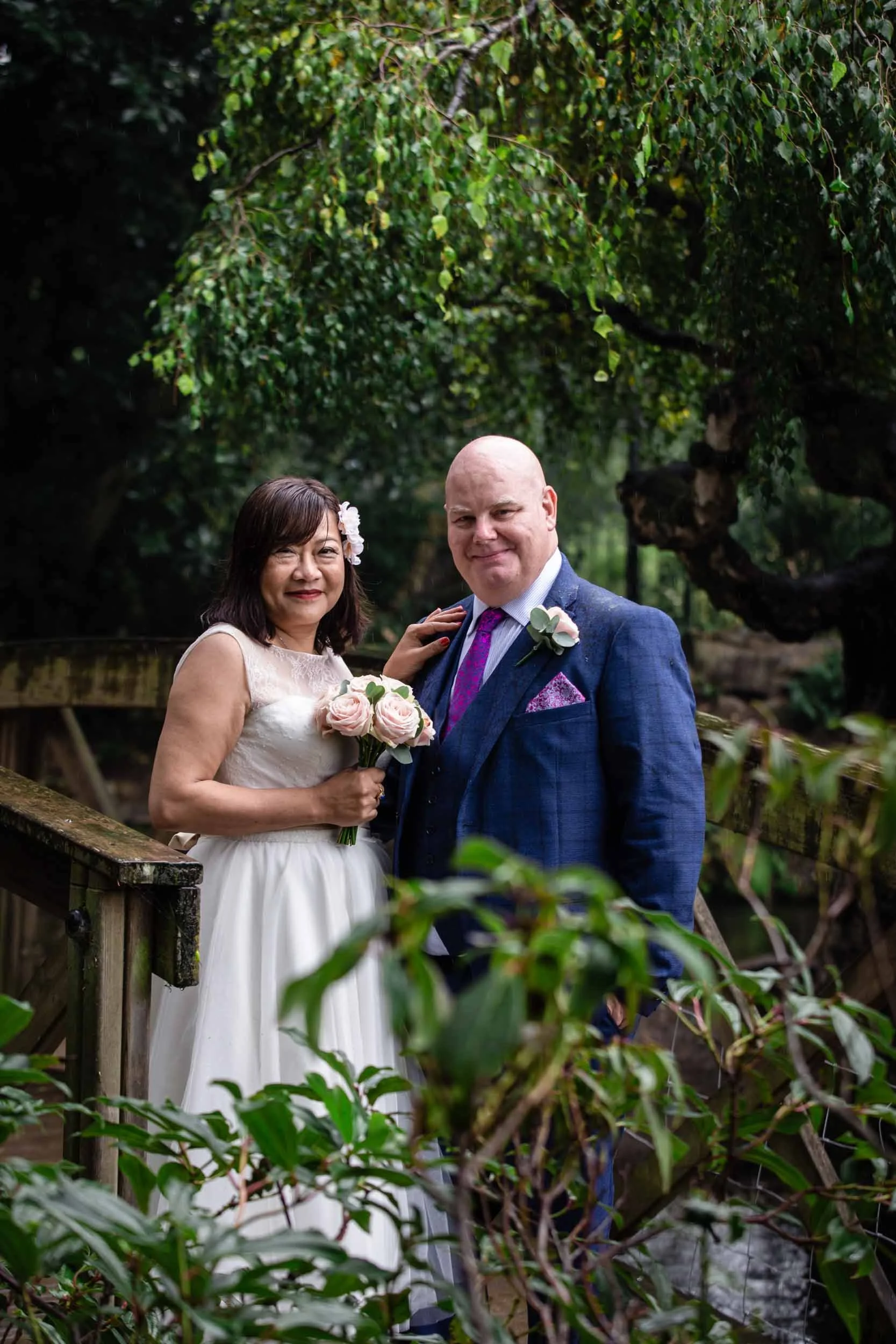 A man and woman in formal wedding attire standing on a wooden bridge surrounded by lush greenery. The woman wears a white wedding dress and holds a bouquet of pink and white roses, while the man wears a blue suit with a purple tie and a white boutonniere. They are smiling and looking at the camera.