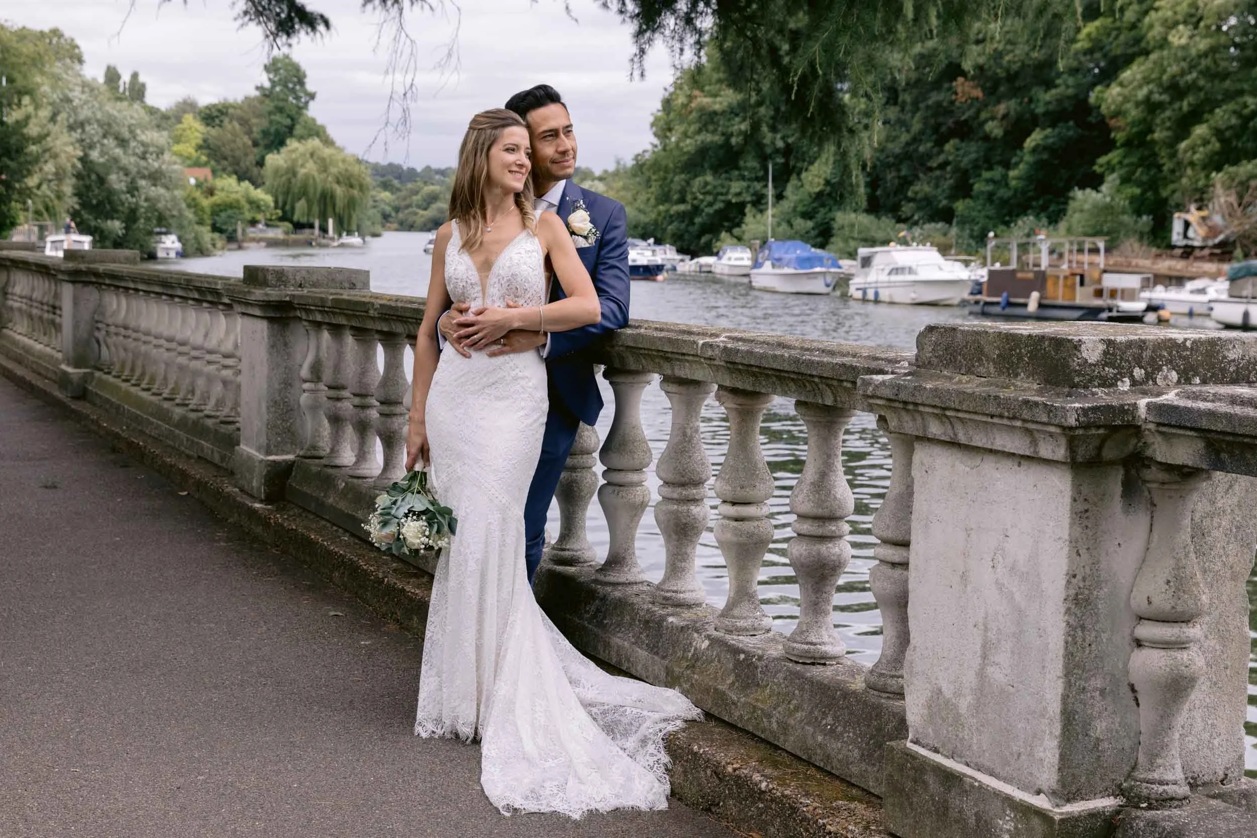 A bride and groom standing by a stone railing near a river with boats, surrounded by lush green trees.