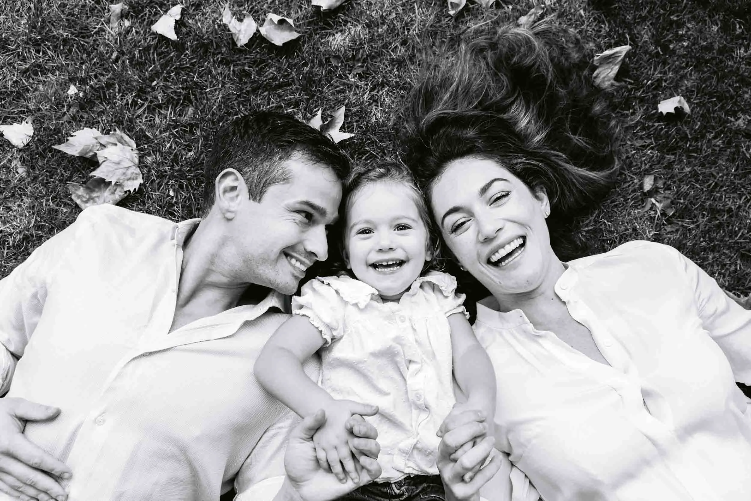 A happy family of three lying on grass outdoors, smiling and looking at the camera. The family includes a man, a woman, and a young girl. They are surrounded by scattered leaves.